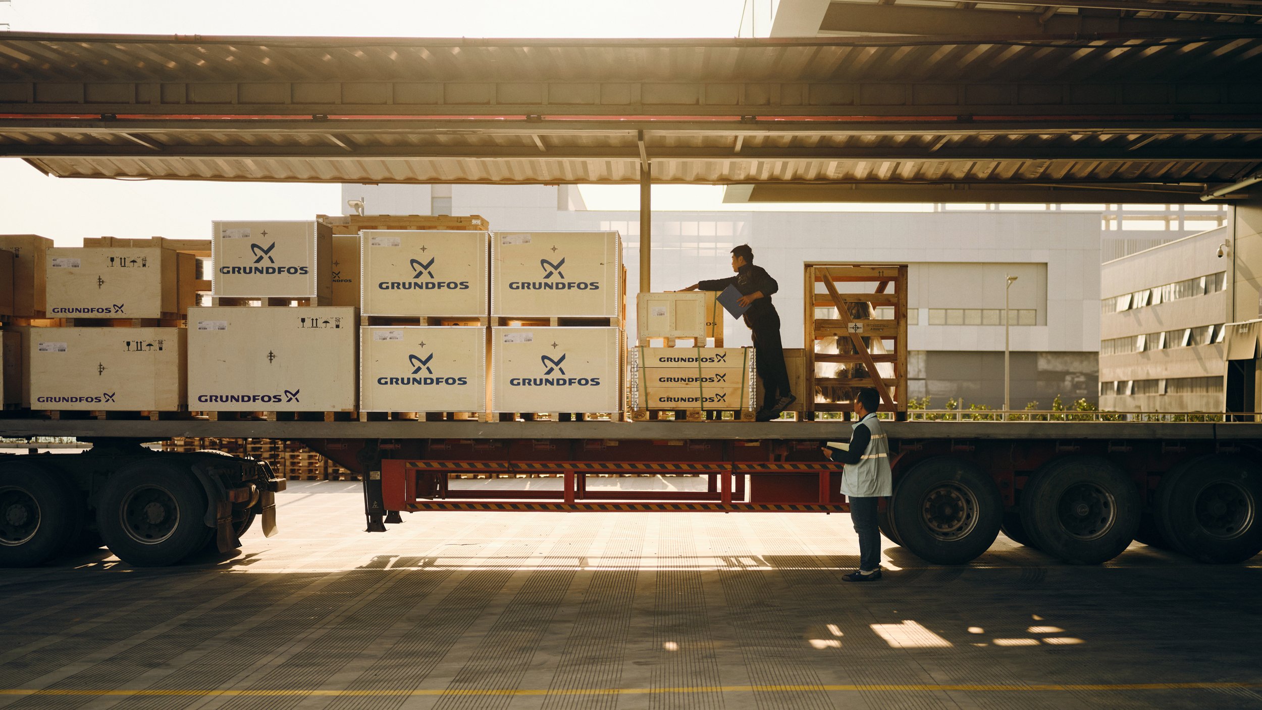 A truck parked at a loading dock with workers unloading boxes labeled 'Grundfos' from the truck.