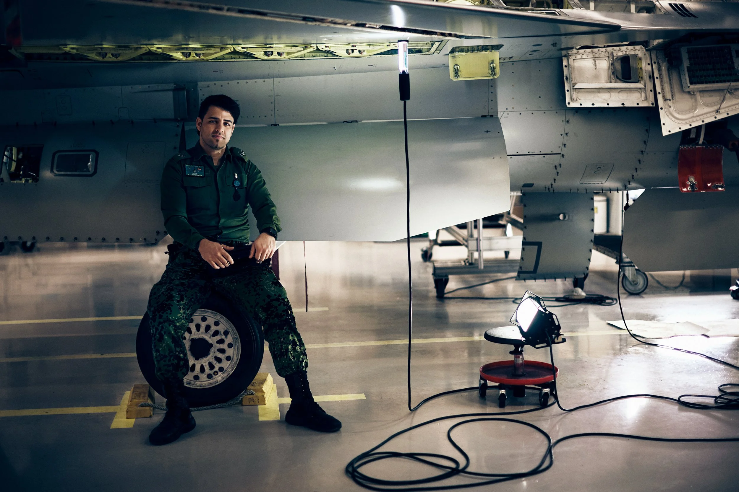Fighter jet mechanic sitting on wheel next to military aircraft in hangar with equipment and cables