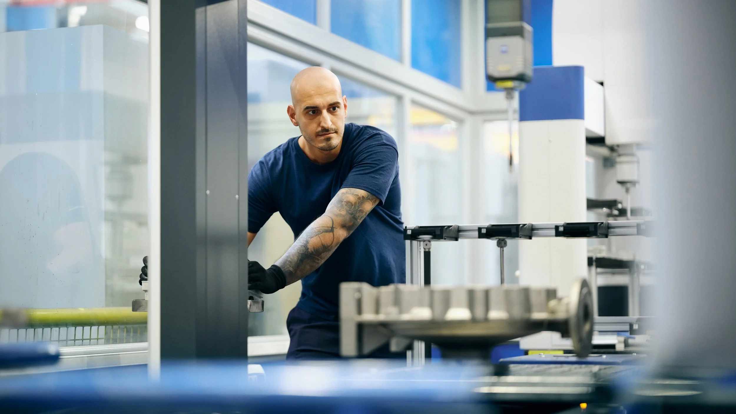 A man with a shaved head and tattooed arm working in a laboratory or manufacturing facility. He is wearing a navy blue shirt and black gloves, and is focused on a task involving machinery.