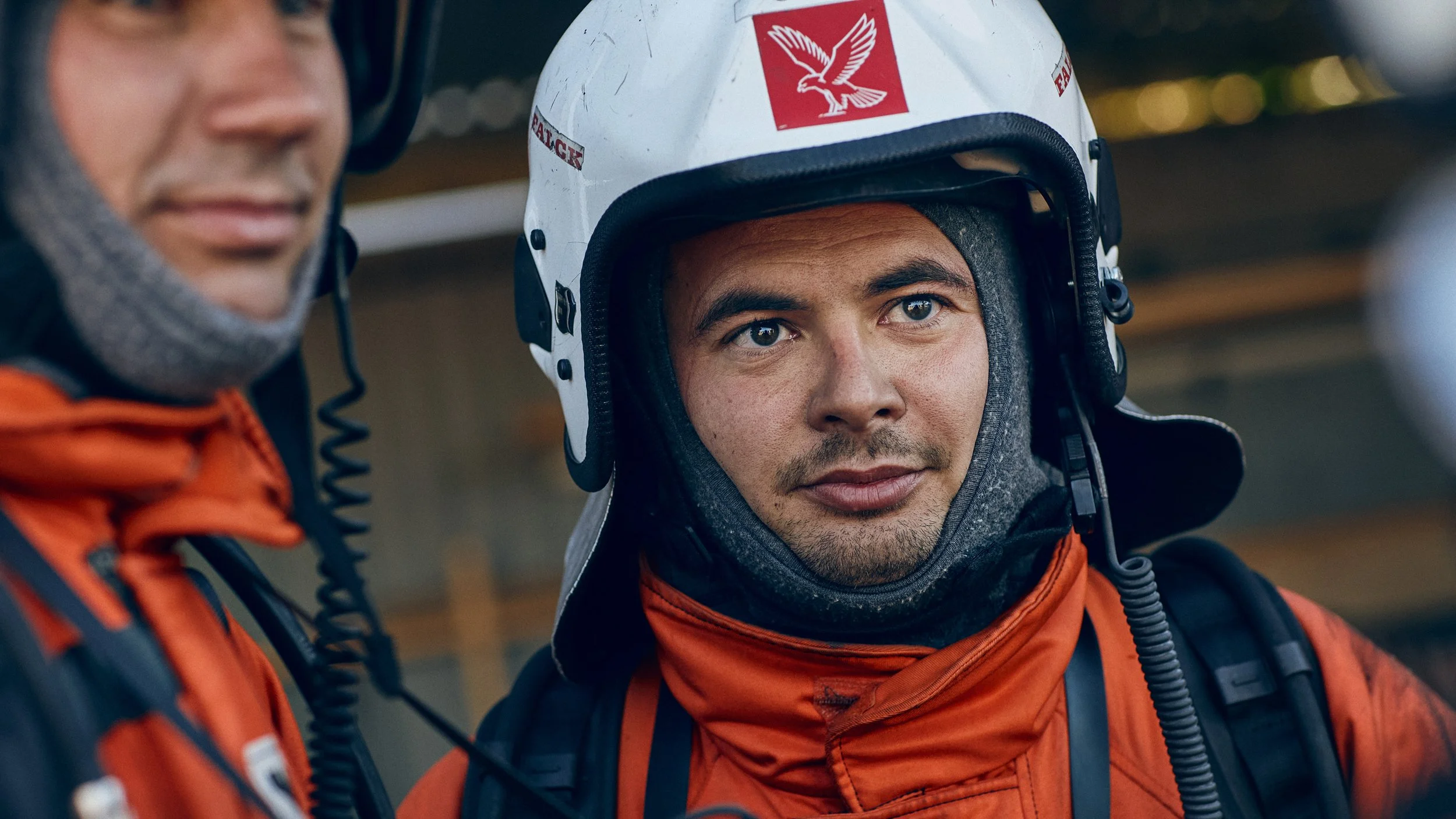 Two rescue workers in orange jackets and helmets, one of whom faces the camera with a serious expression, the other partially visible.