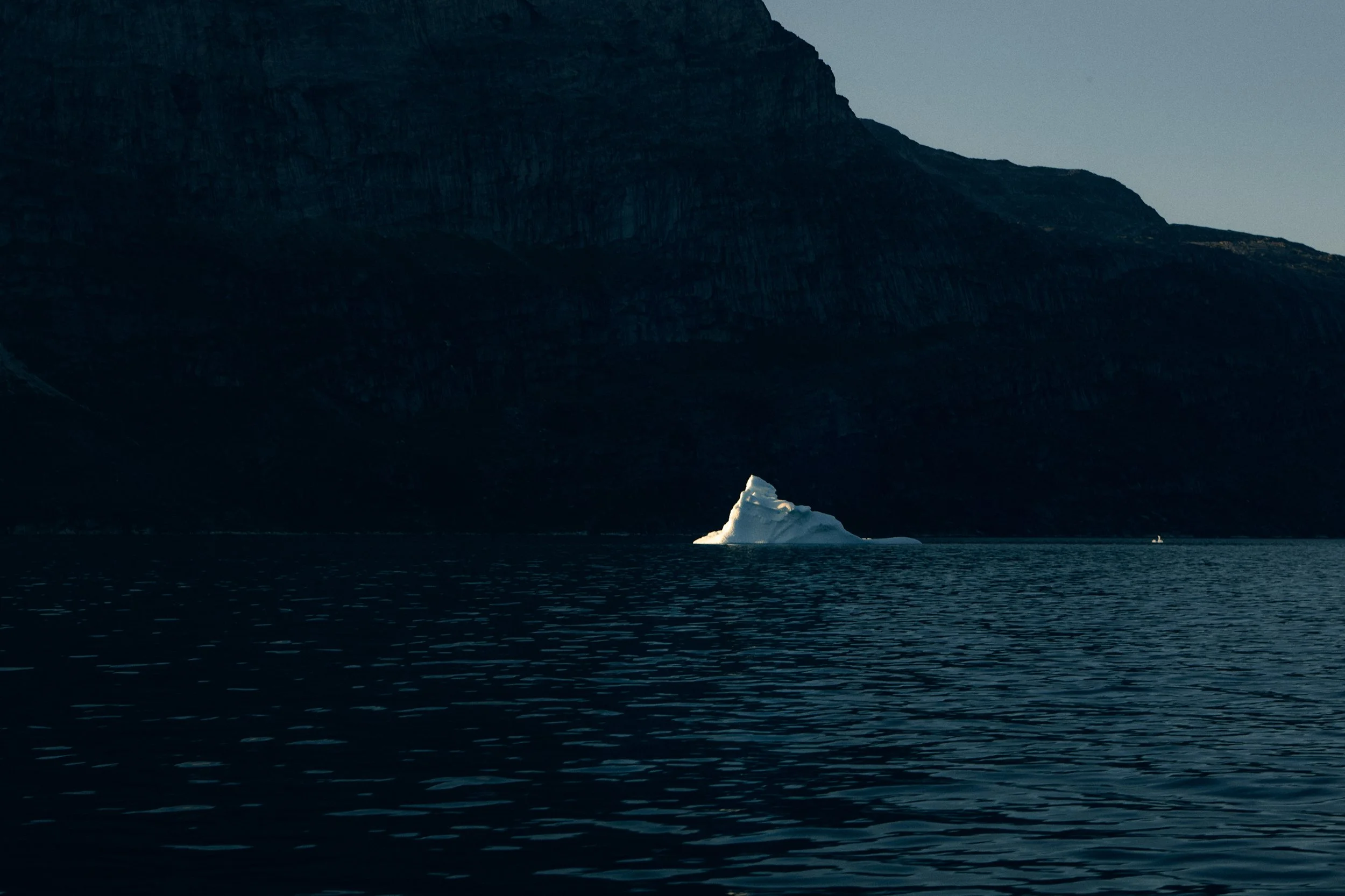 Iceberg floating in dark water beneath steep cliffs, quiet arctic landscape