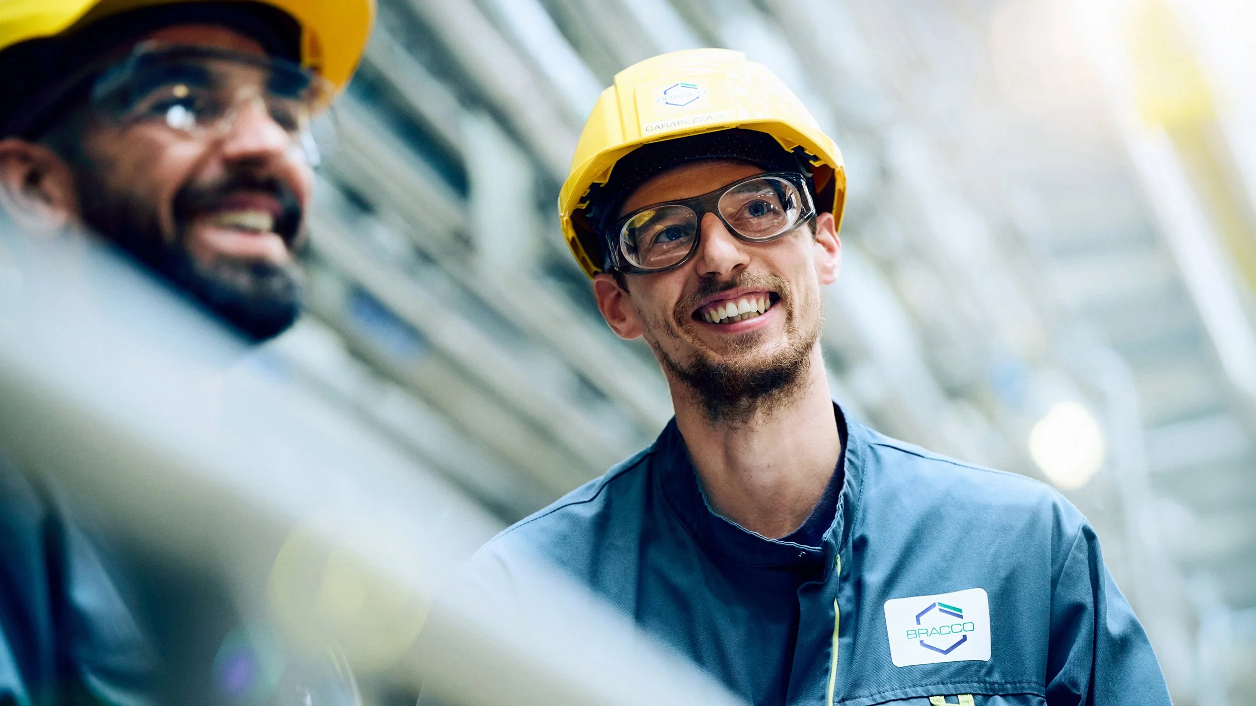 Two industrial workers wearing yellow safety helmets and safety glasses, smiling while working together in an industrial setting.