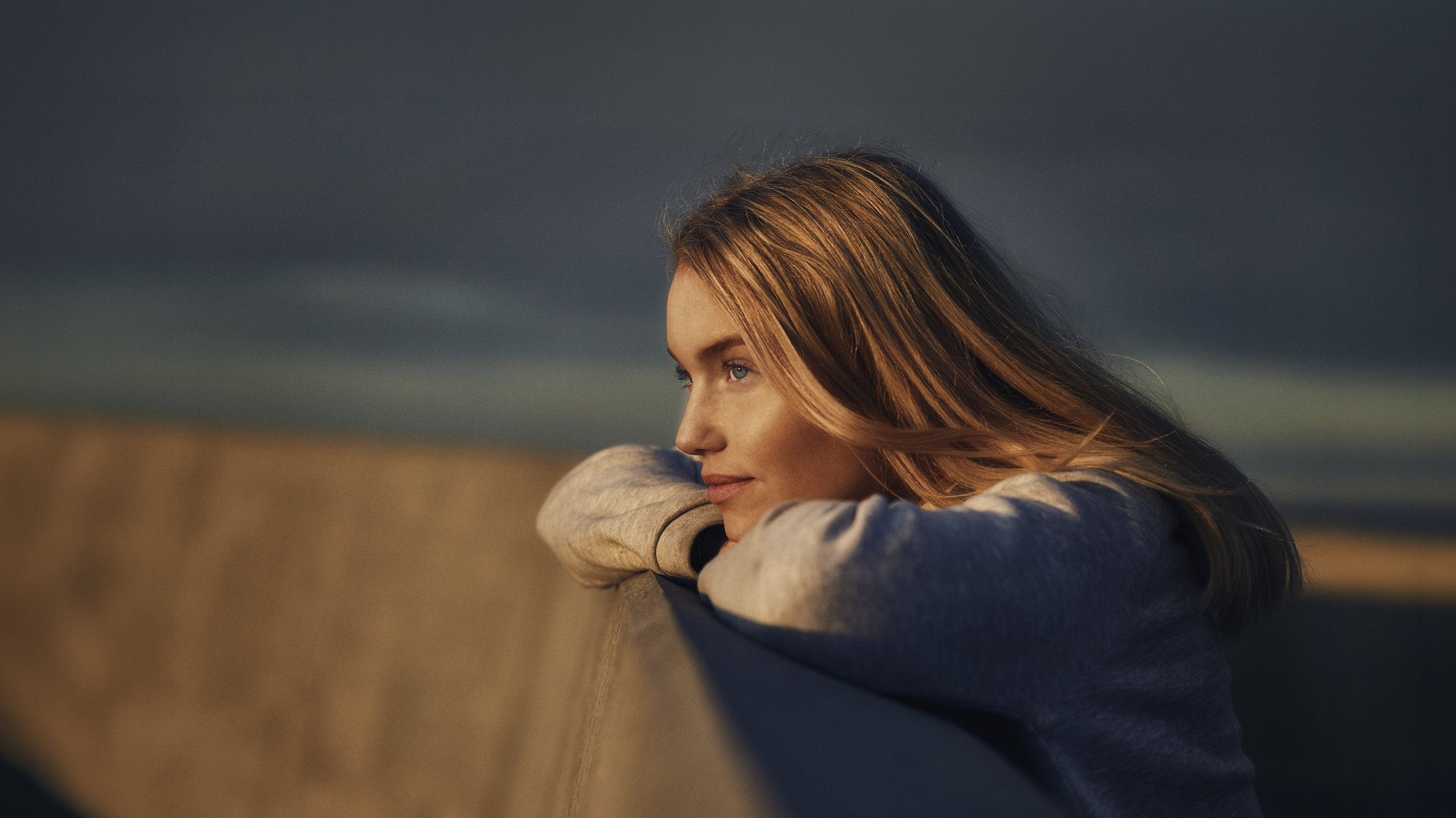 A young woman with long, reddish-brown hair leans on her arm, looking thoughtfully into the distance against a dark, cloudy sky at dusk or dawn.