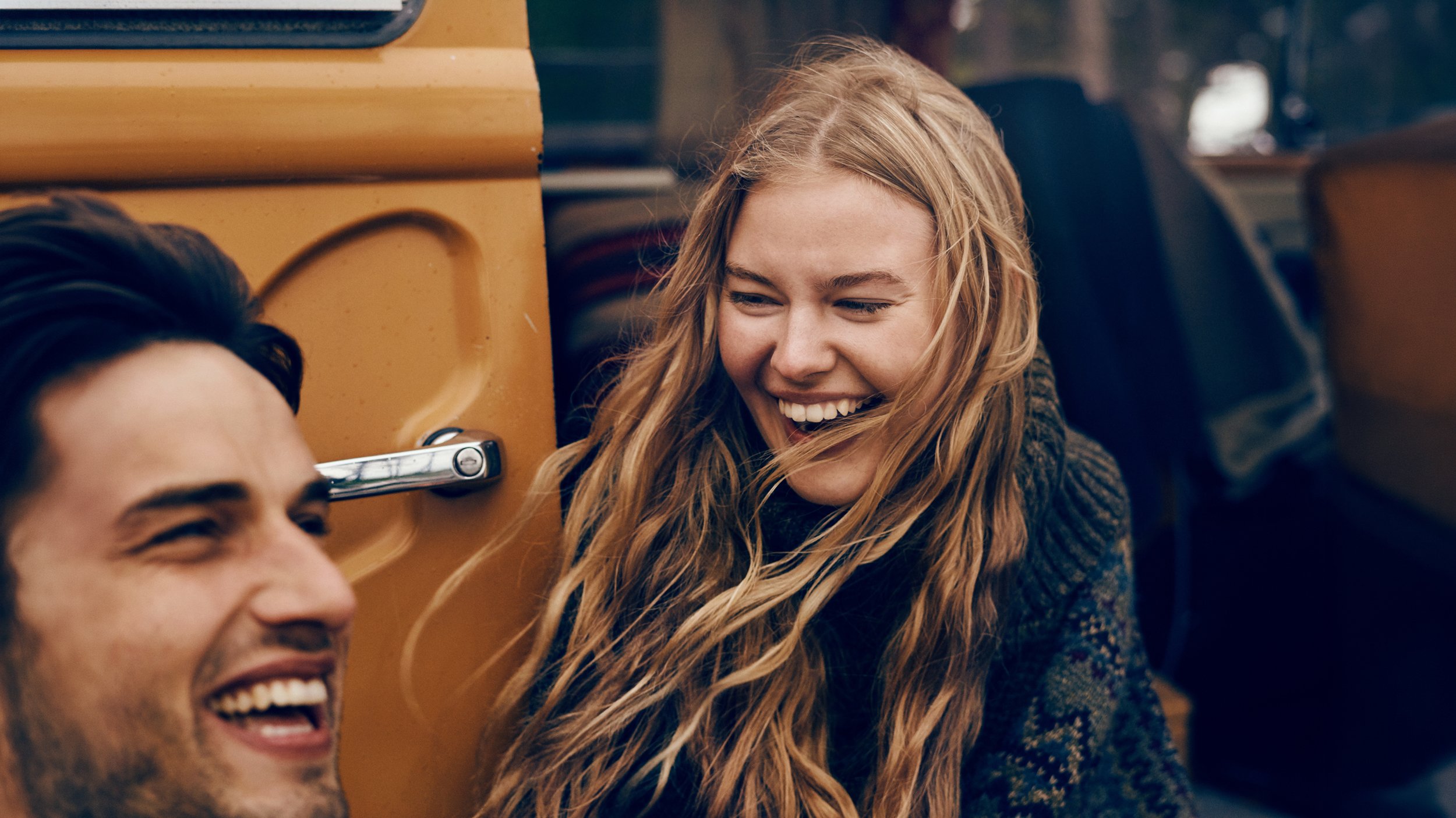 Two friends, a woman with long red hair and a man with dark hair, smiling and laughing together outside near a yellow vehicle.