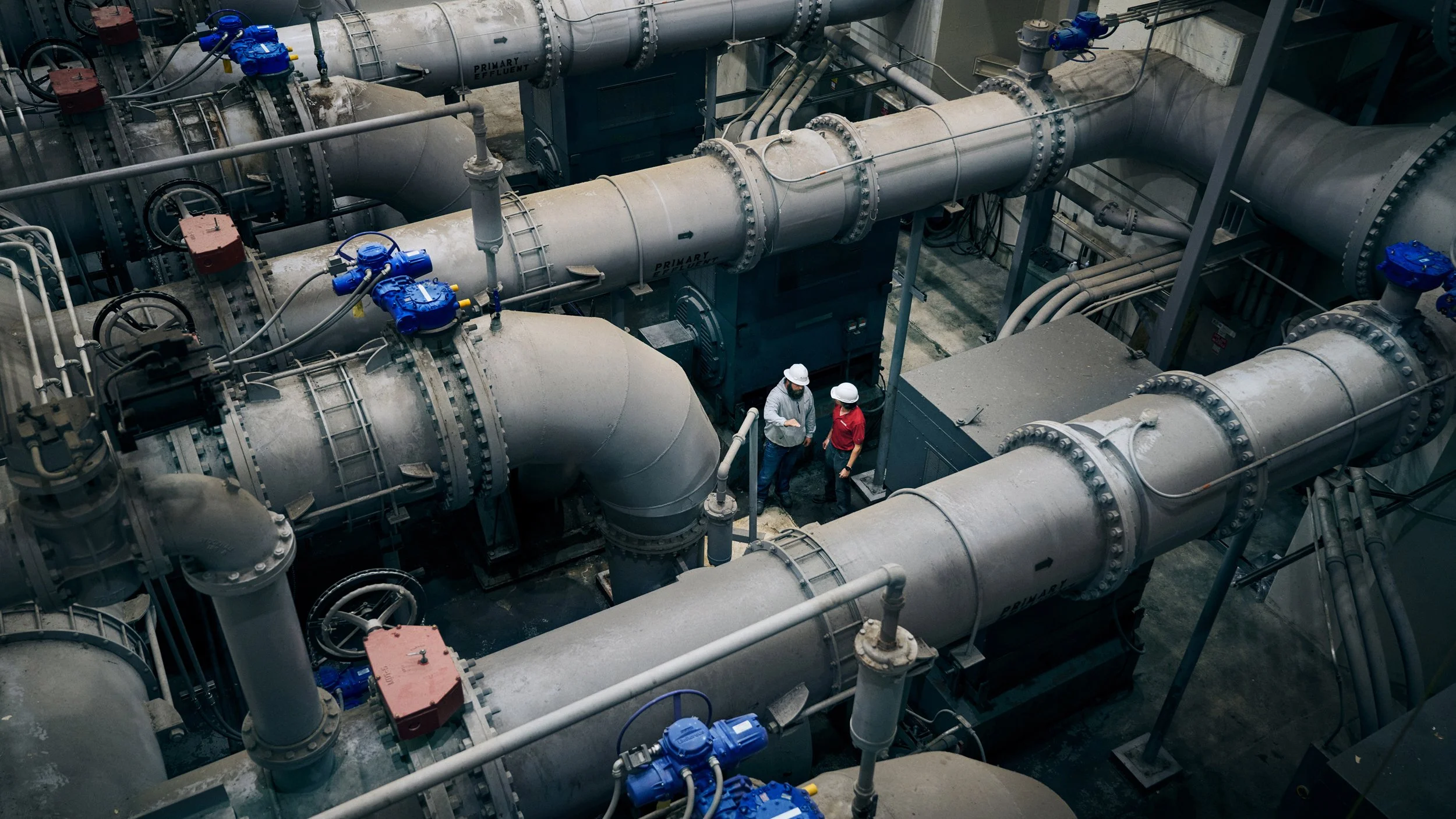 Two workers wearing safety helmets and protective gear standing among large industrial pipelines and valves inside a factory or processing plant.