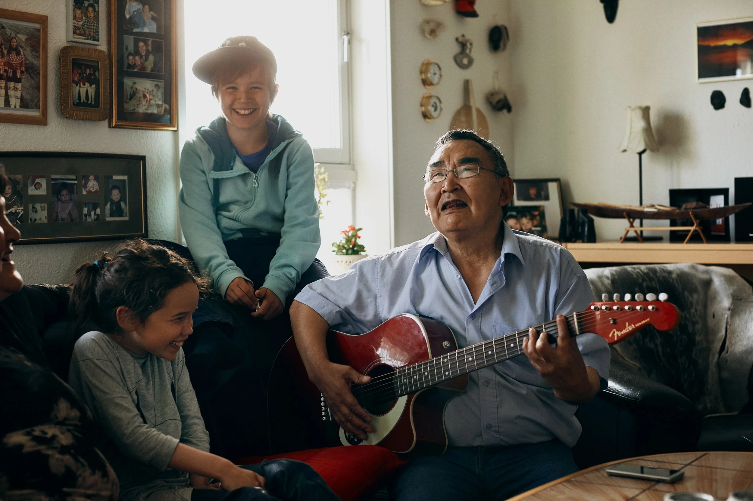 Older man playing guitar surrounded by children, shared moment in living room