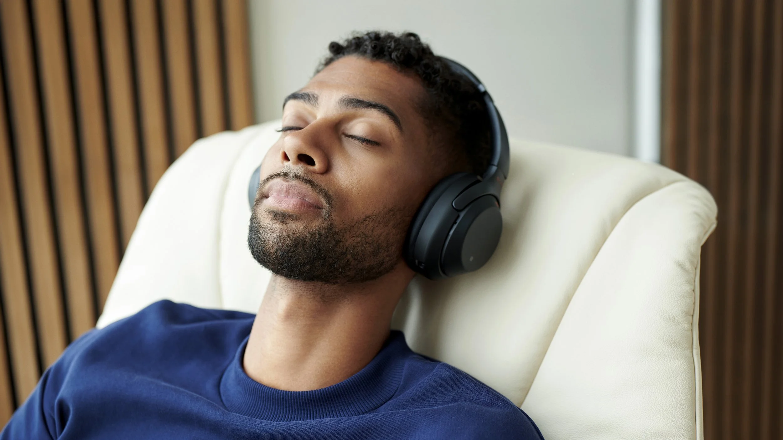 Man with curly hair and beard wearing headphones, resting with eyes closed on a cream-colored chair in a room with wooden panels.