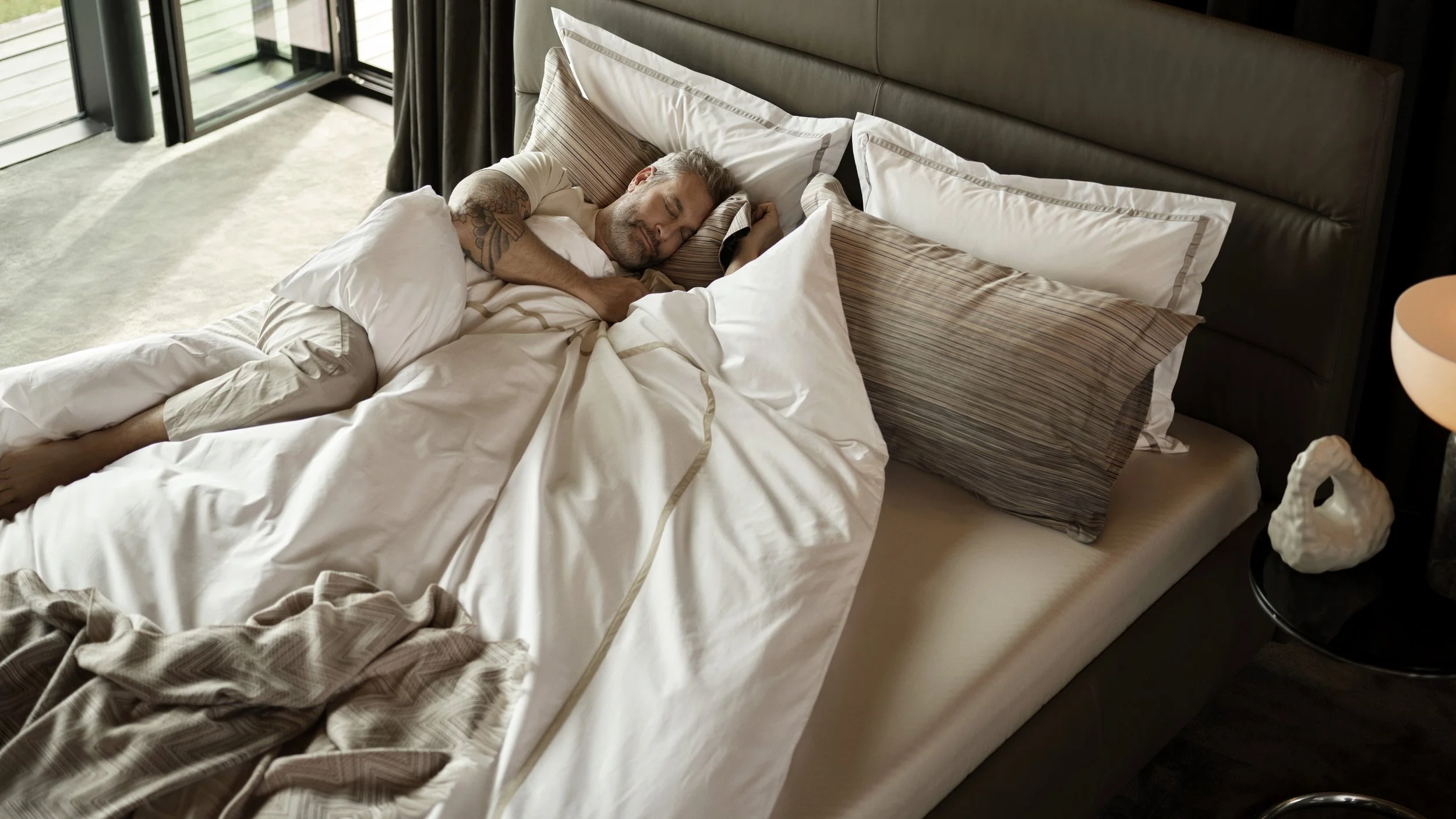 A man with tattoos sleeping peacefully on a bed with white sheets, multiple pillows, and a headboard in a modern hotel room. There is a lamp on a bedside table and a large window nearby.