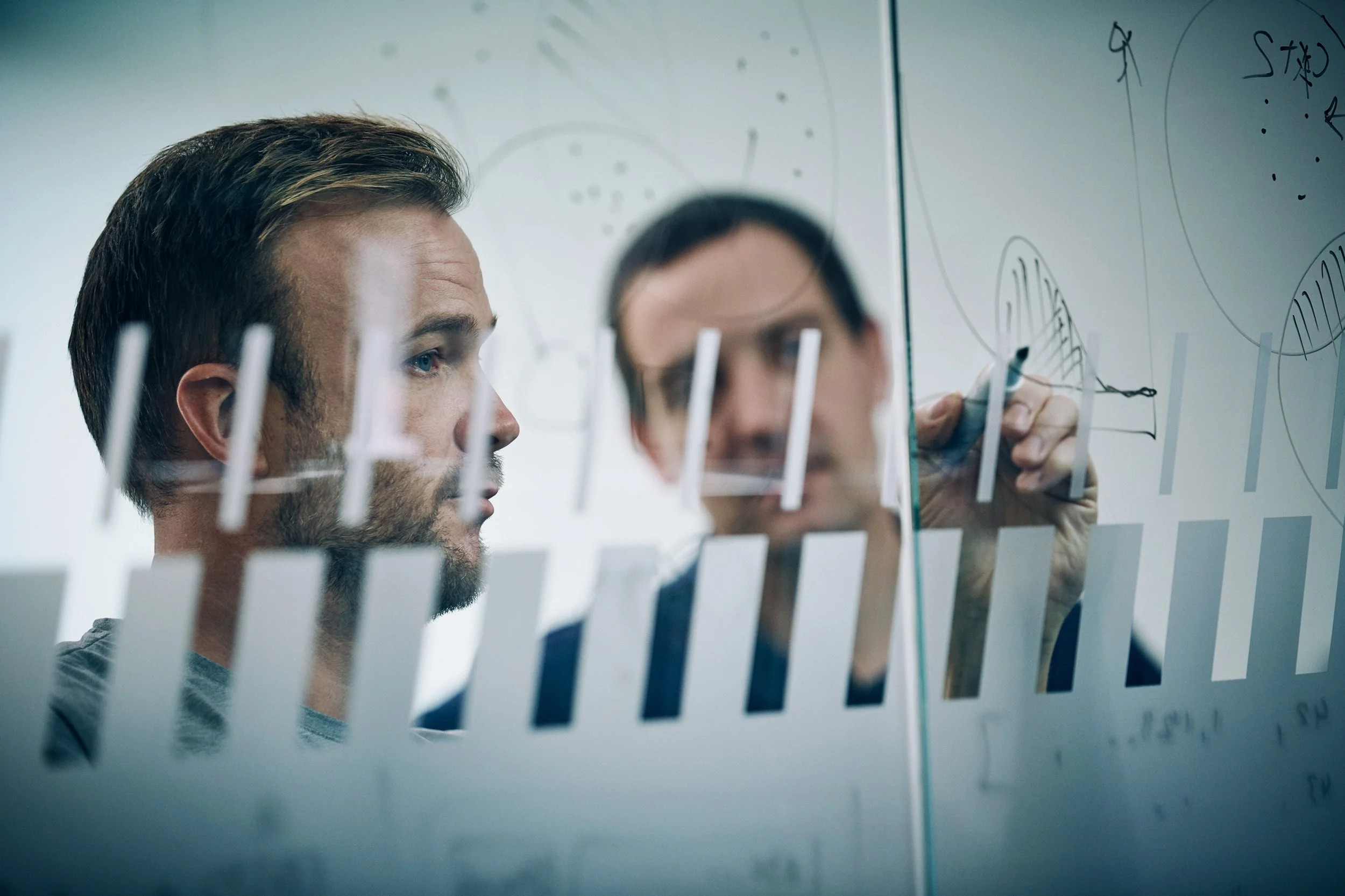 Two men reviewing diagrams on glass board in office
