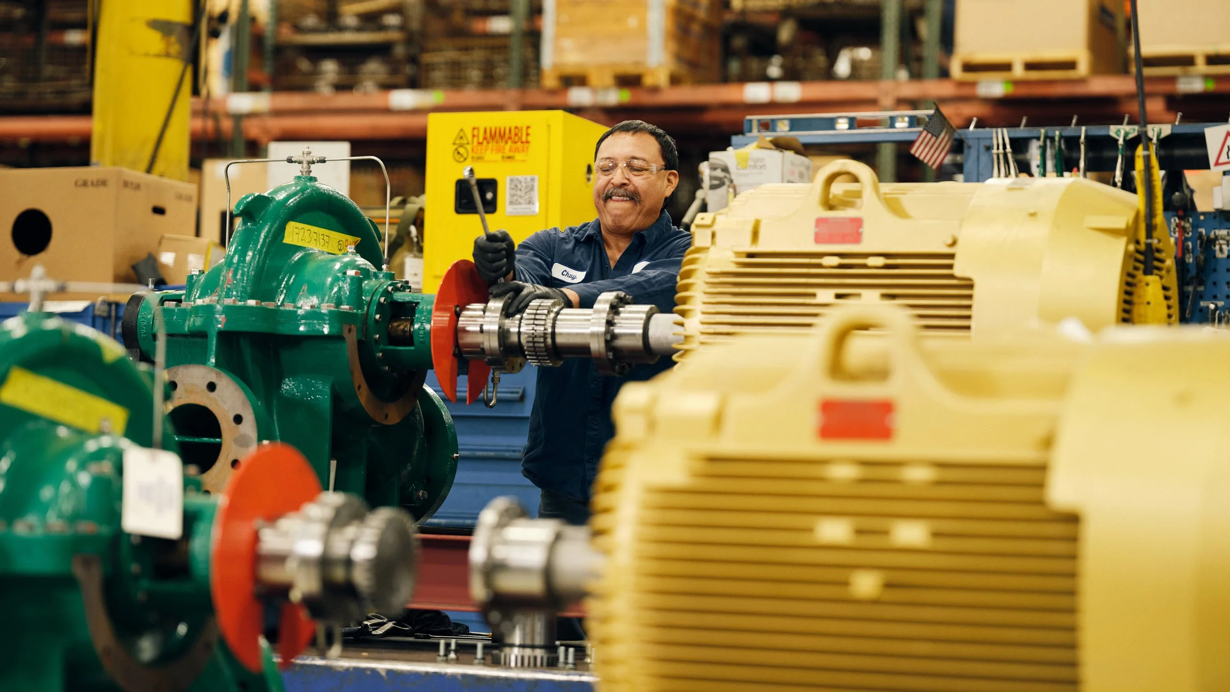 A man in safety glasses and dark work clothes is working with industrial machinery in a warehouse. He is smiling and using a tool on a large green machine, surrounded by various industrial equipment and yellow storage containers.
