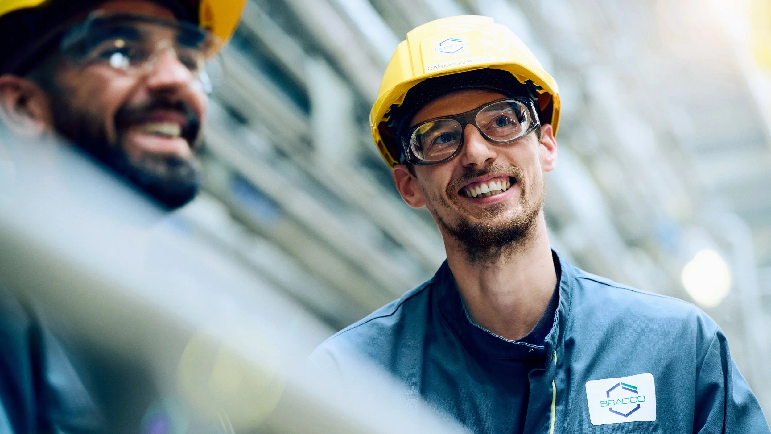 Two workers with safety helmets and glasses in an industrial setting, one smiling and the other slightly blurred.