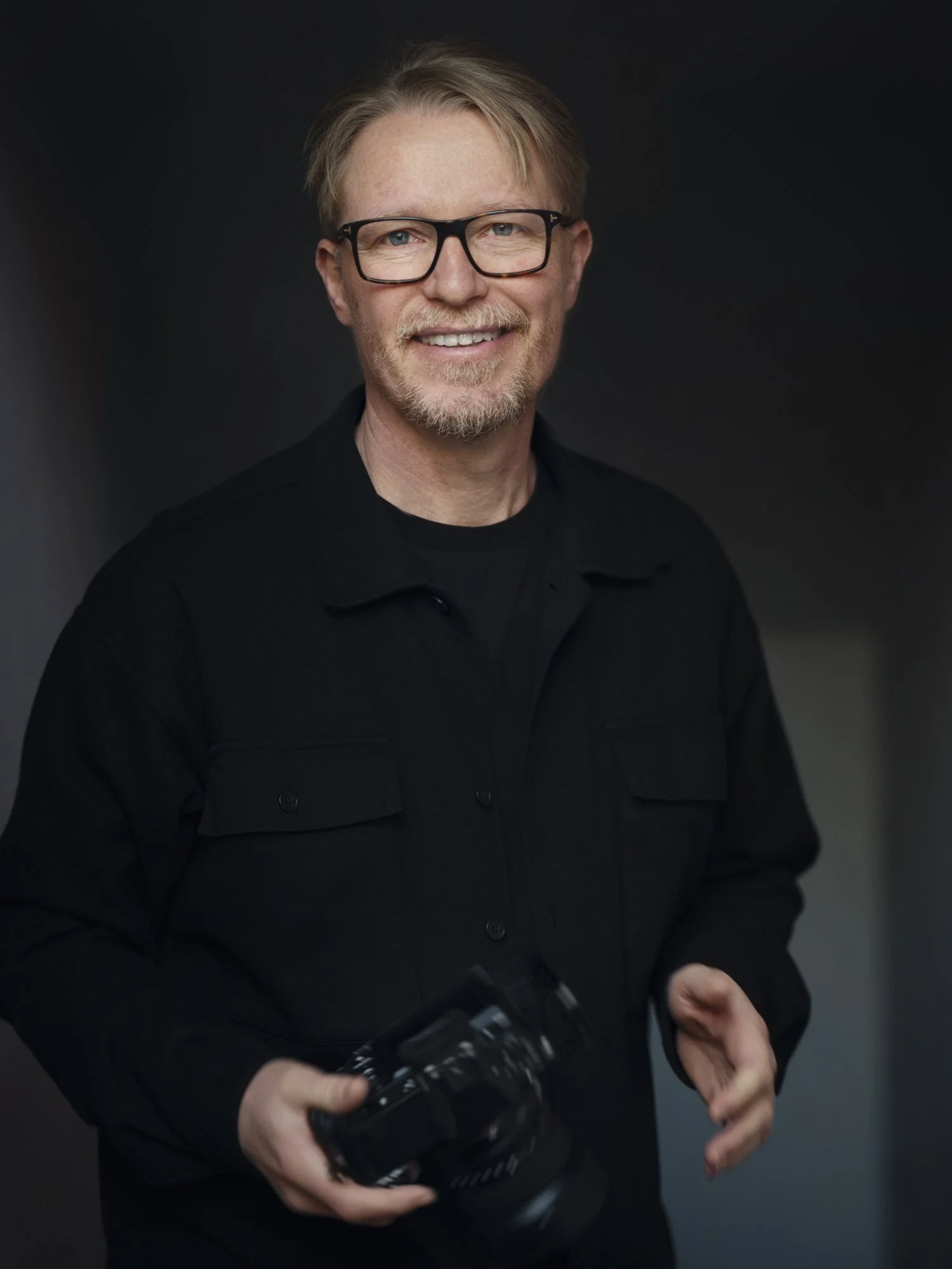 A man with blond hair, glasses, and a beard smiling while holding a camera, against a dark background.