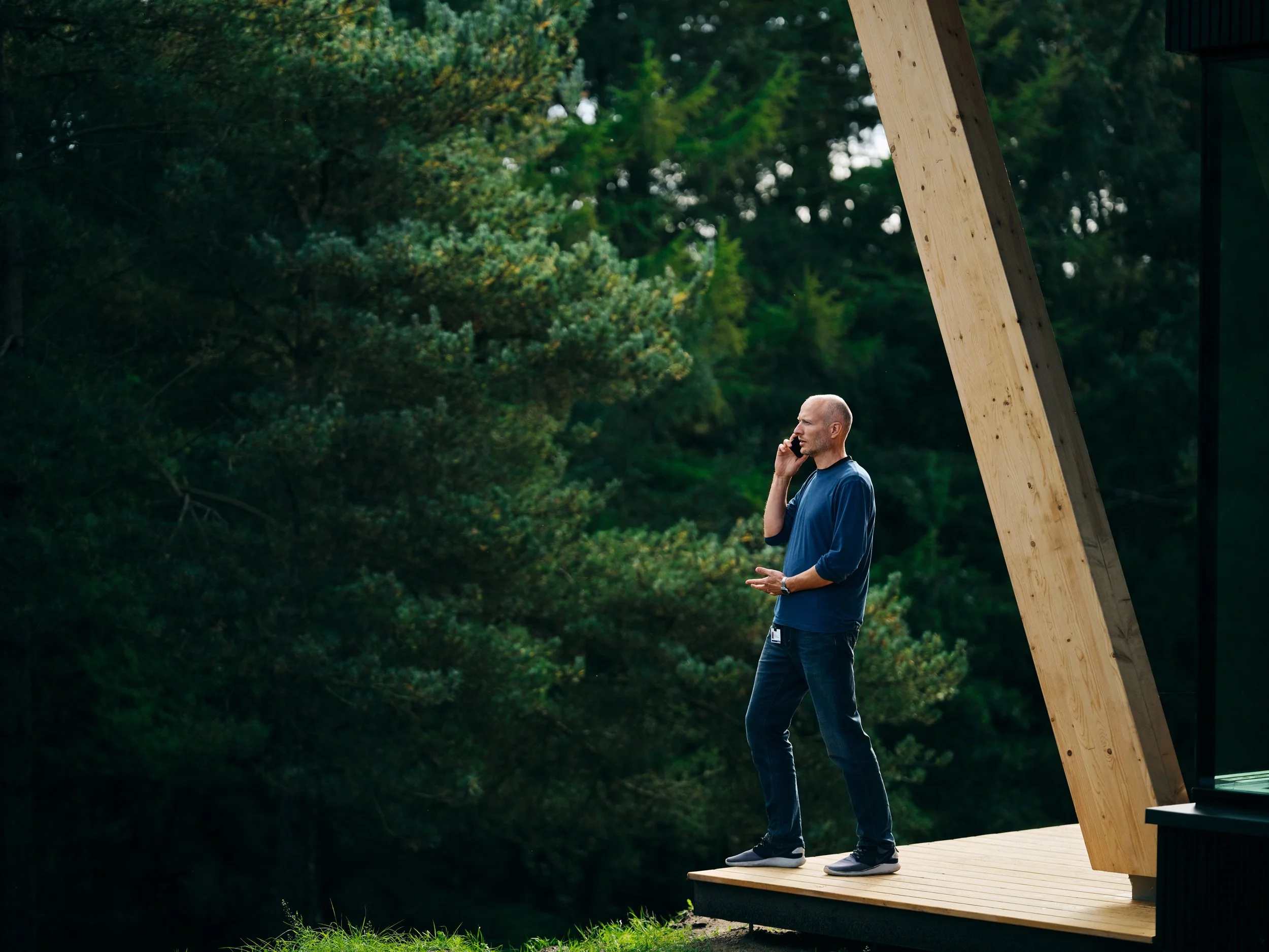 Man standing on modern wooden terrace outside building, speaking on phone with forest landscape in background