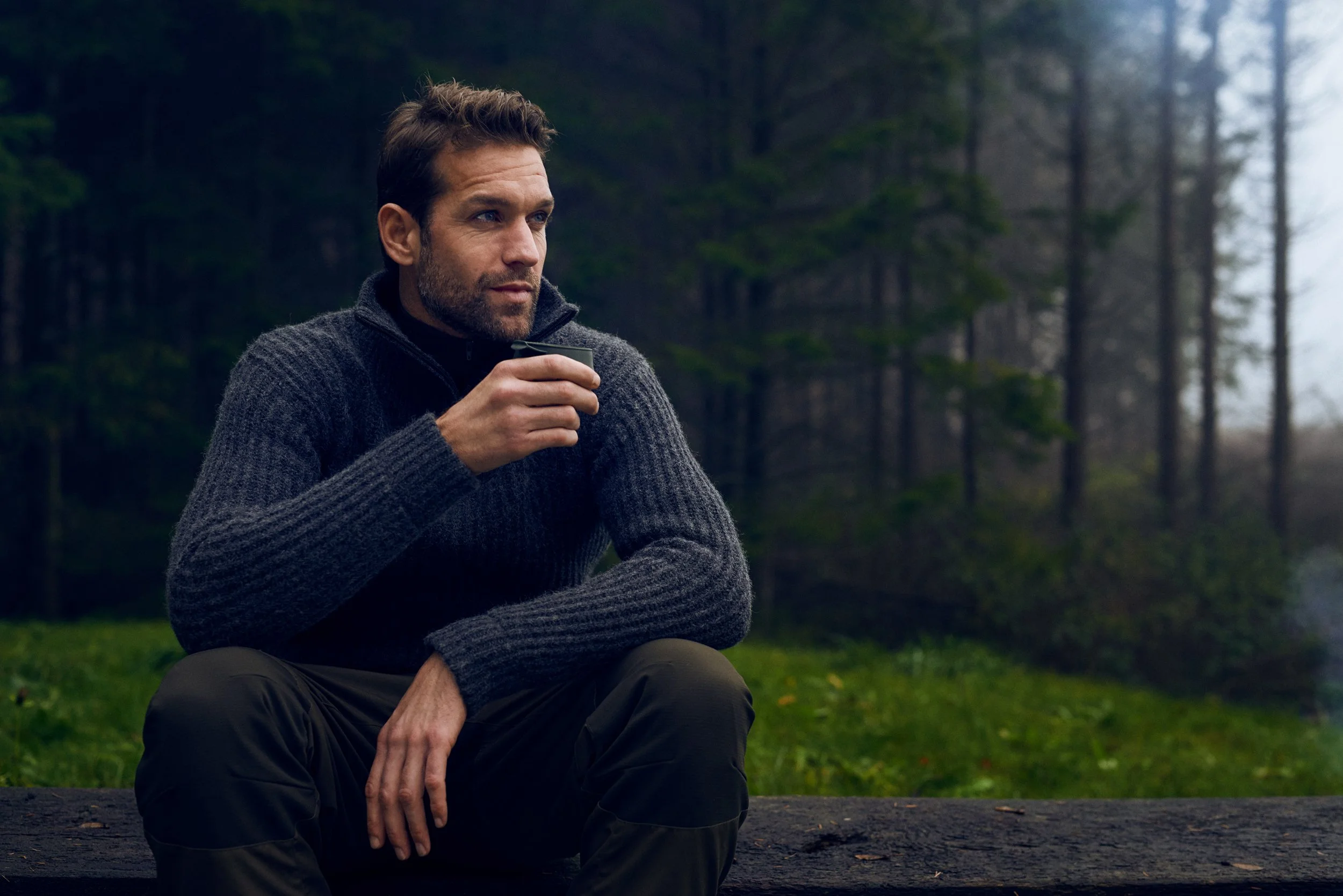 Man sitting on log holding cup in forest, quiet pause in outdoor setting
