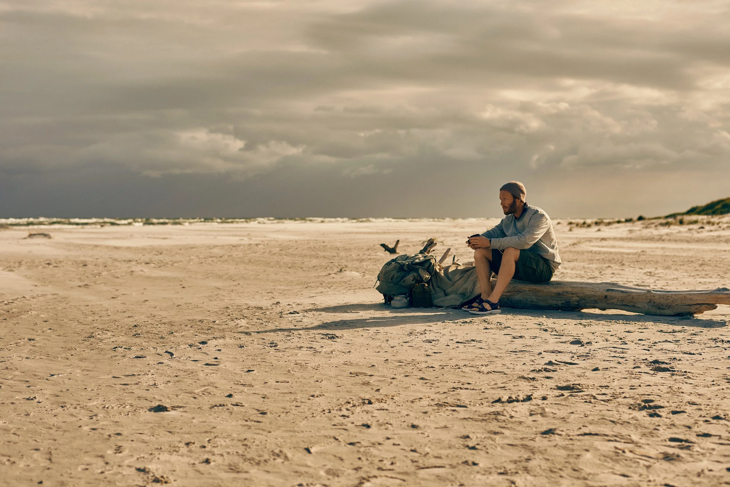Man sitting on driftwood on beach with backpack, coastal outdoor moment