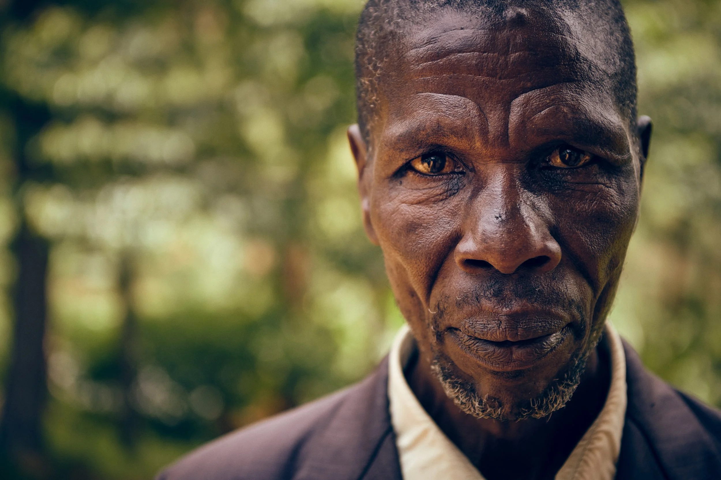 Portrait of older man standing outdoors, direct gaze against forest backdrop
