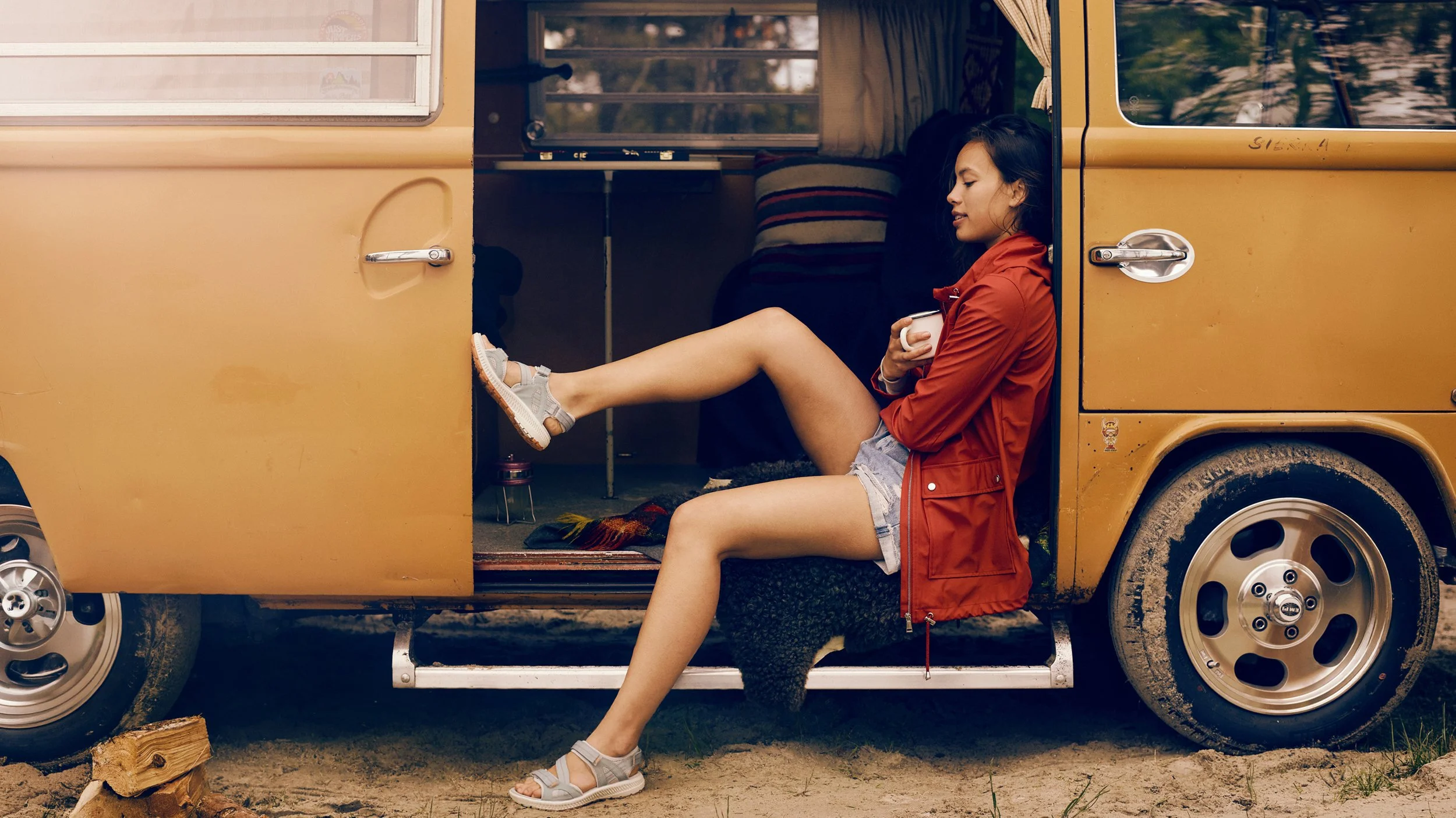 A young woman is sitting in the open doorway of a yellow vintage van, holding a white mug, with her legs stretched out and wearing casual summer clothes and sandals.