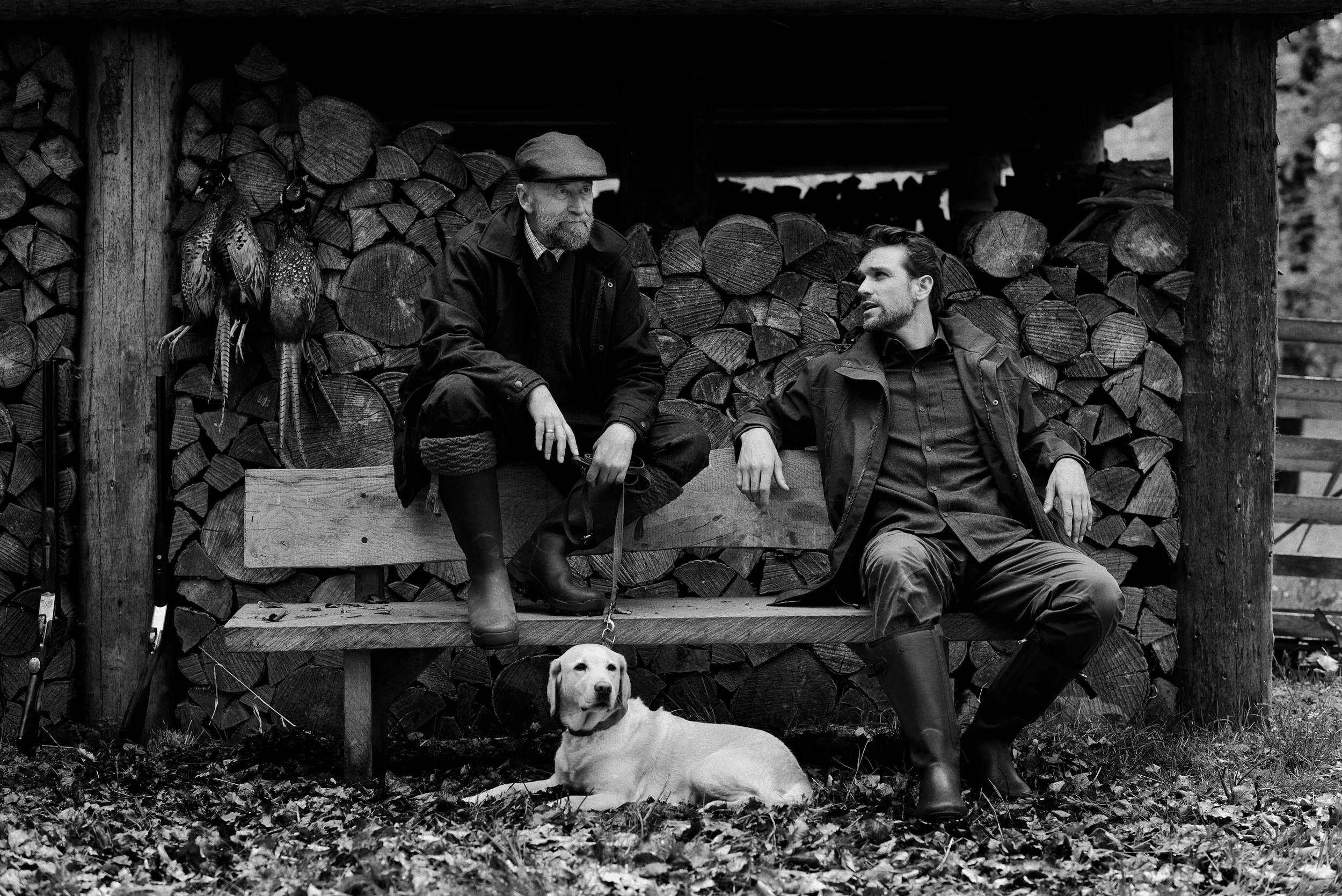 Two men seated together with dog in front of wood stack, quiet conversation in traditional setting
