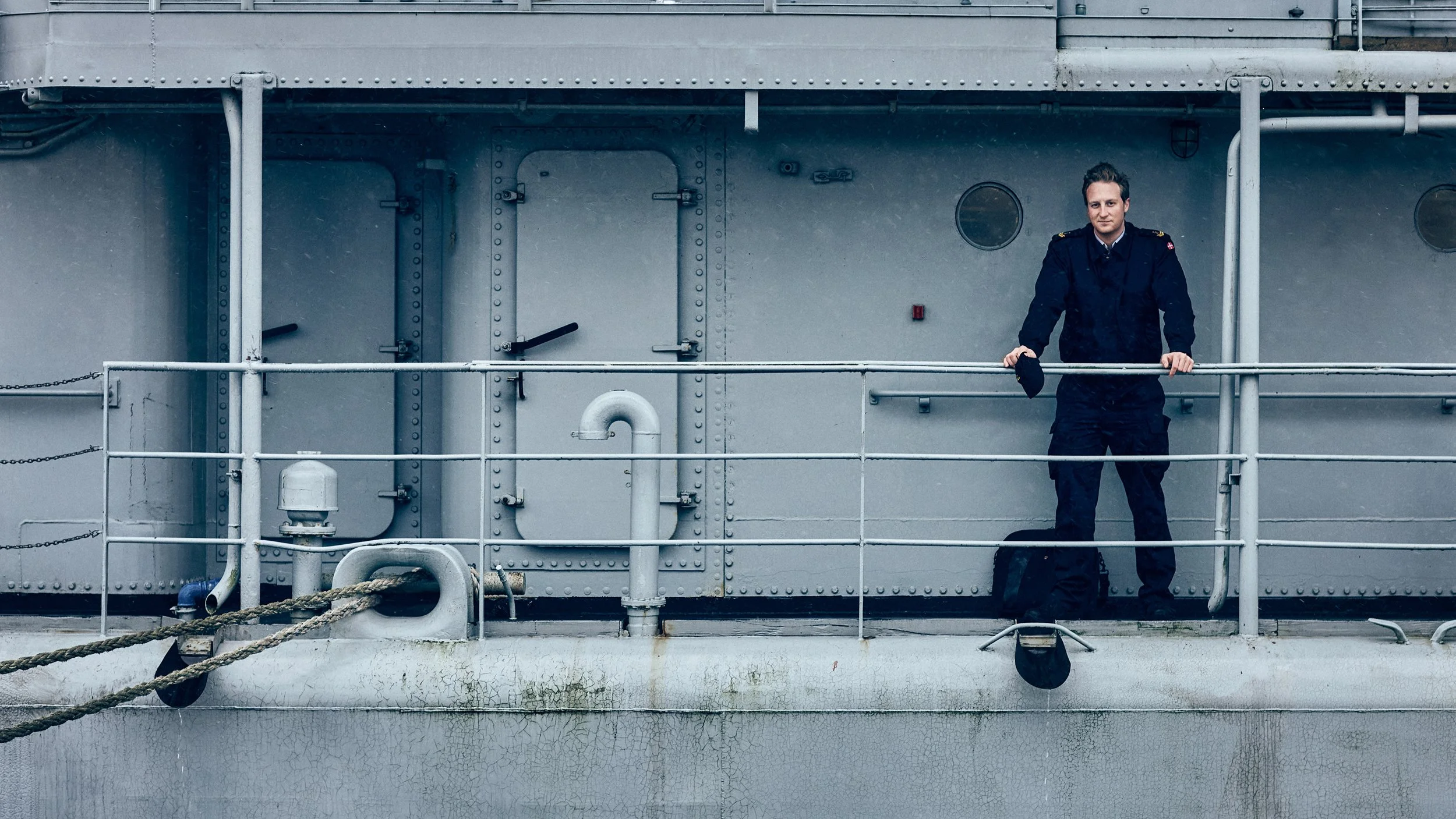 A man in a navy uniform with gold epaulets standing on the deck of a ship, holding a black cap in his right hand, with gray industrial ship structures and railings in the background.