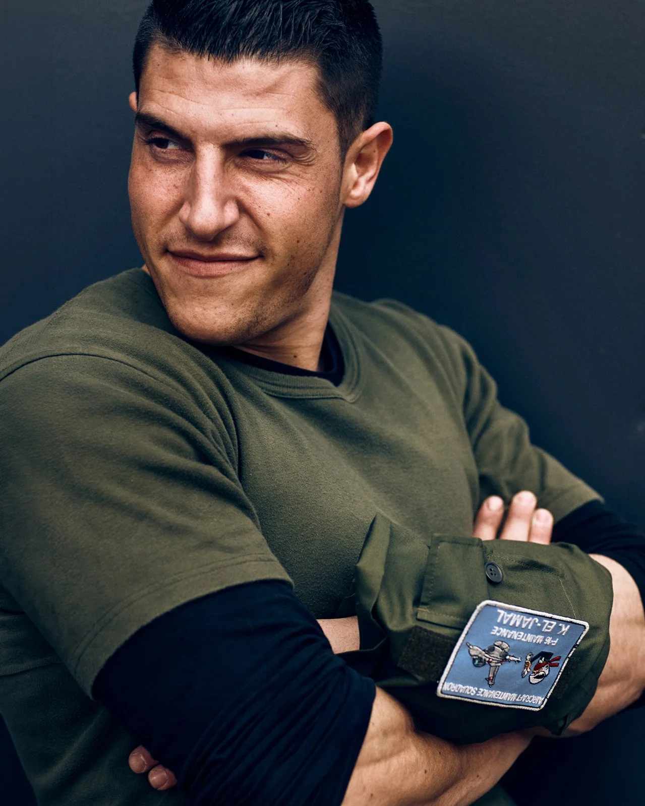 A young man with short dark hair and wearing a green military-style uniform with patches, sitting with arms crossed against a dark background.