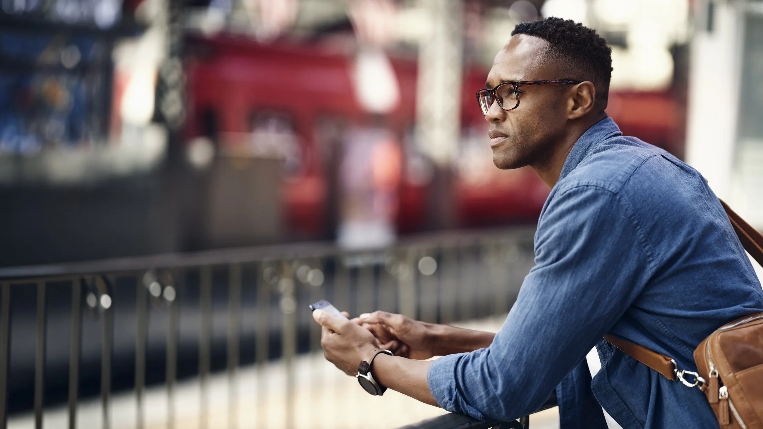 A young man with dark skin, black glasses, and short curly hair sitting outdoors, looking to the right while holding a smartphone in his hands. He is wearing a denim shirt and a black watch, with a brown backpack resting on his back. The background i