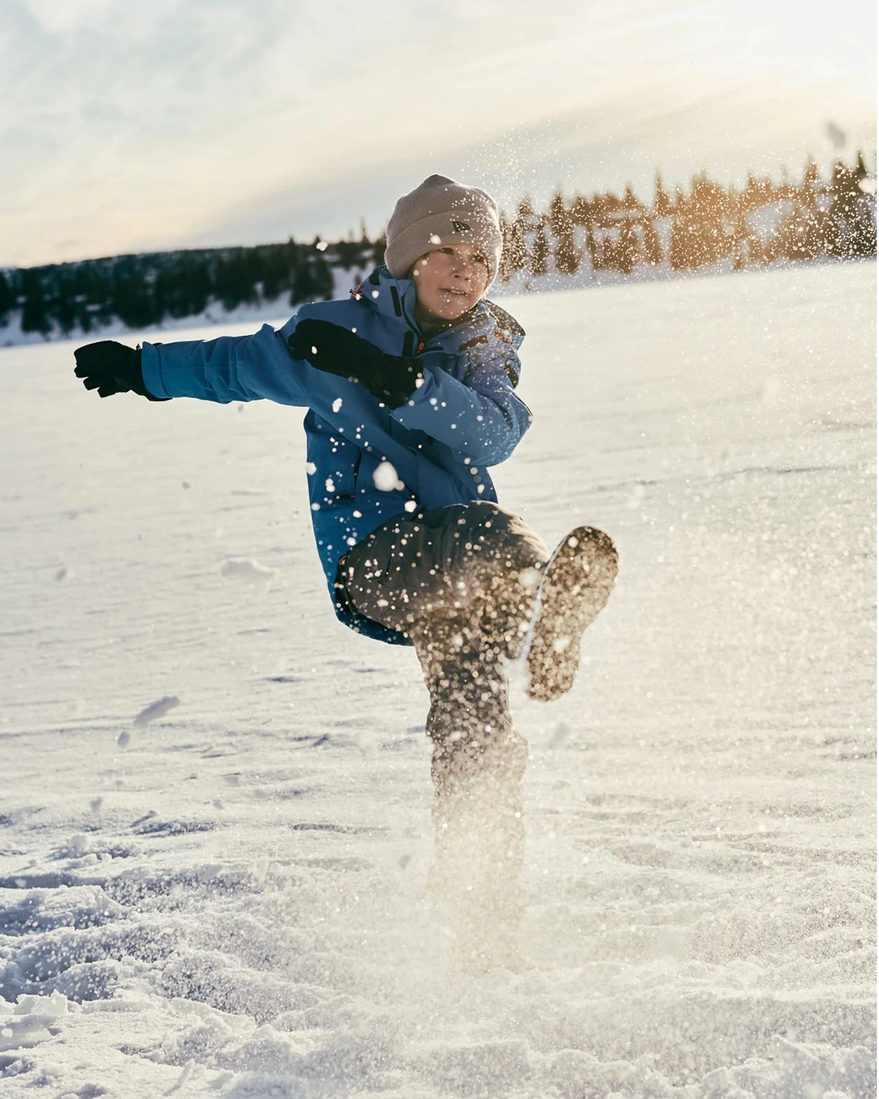 A young boy playing and kicking up snow in a winter landscape during sunset.