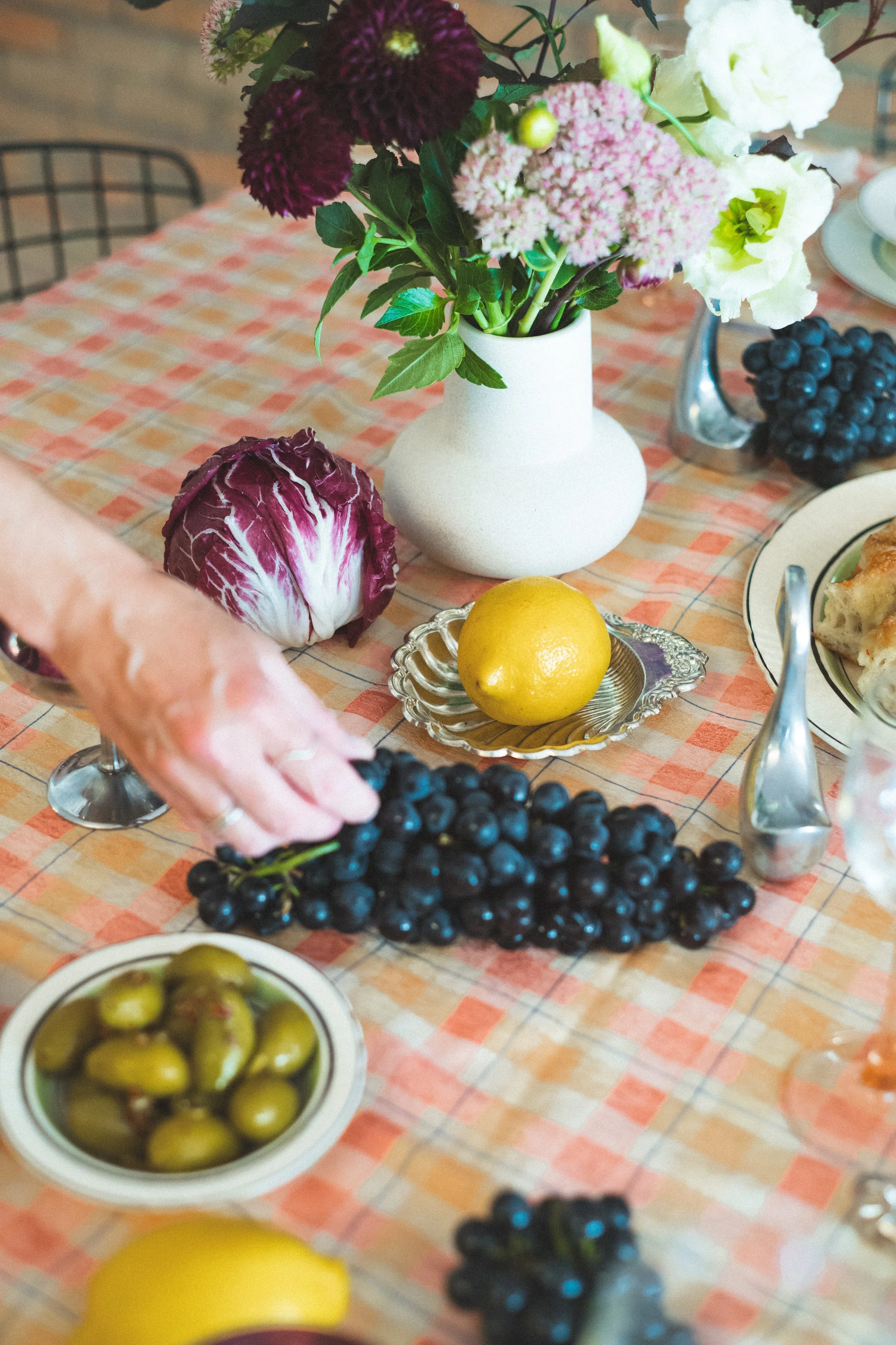 A table with a floral arrangement in a white vase, a head of radicchio, a lemon on a silver dish, green and purple grapes, a bowl of green olives, and some other dishes, with a patterned tablecloth.