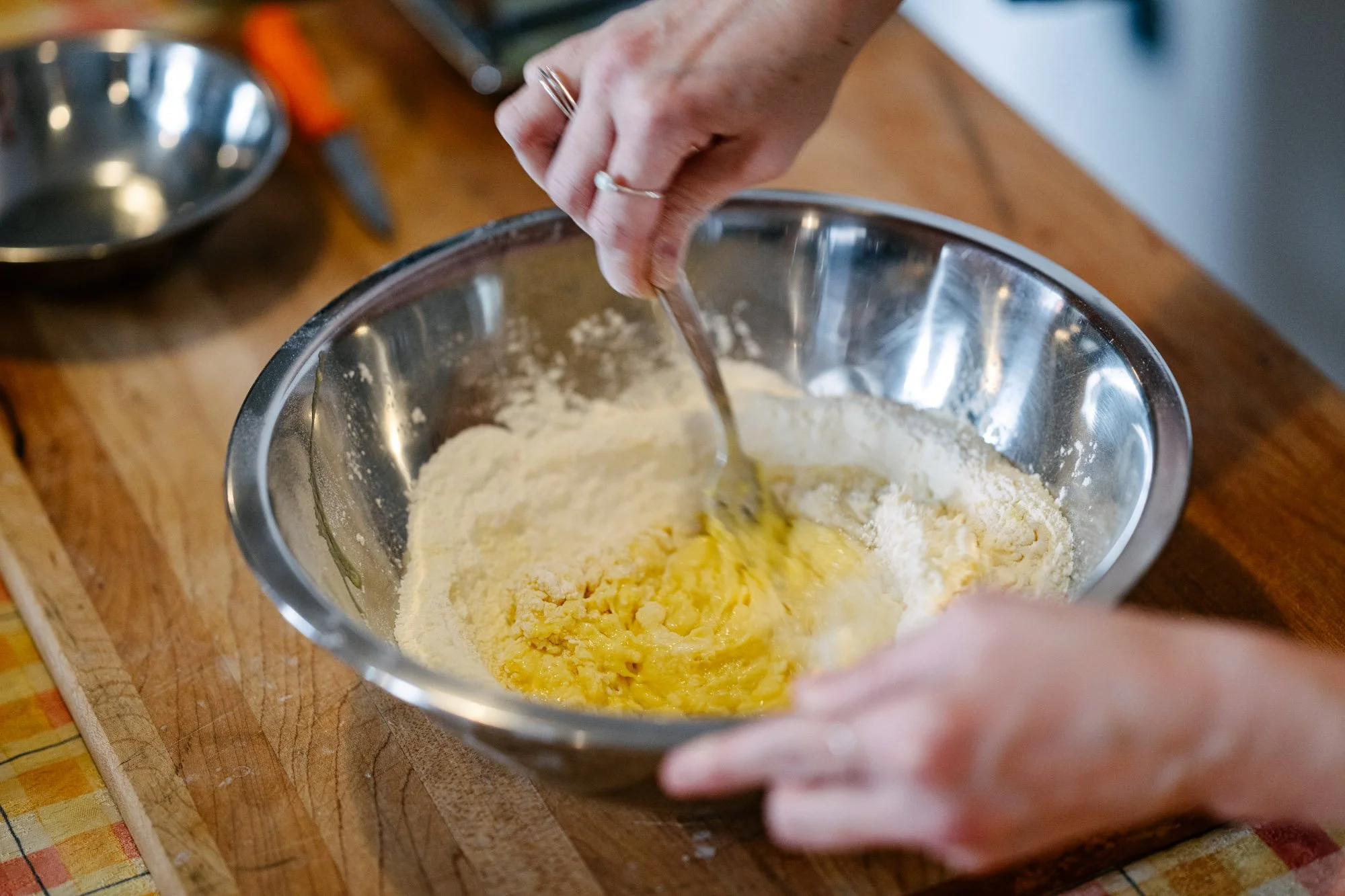 A table set up for a cooking class with stainless steel mixing bowls, wooden cutting boards, and utensils in an industrial-style kitchen with exposed brick walls and large windows.