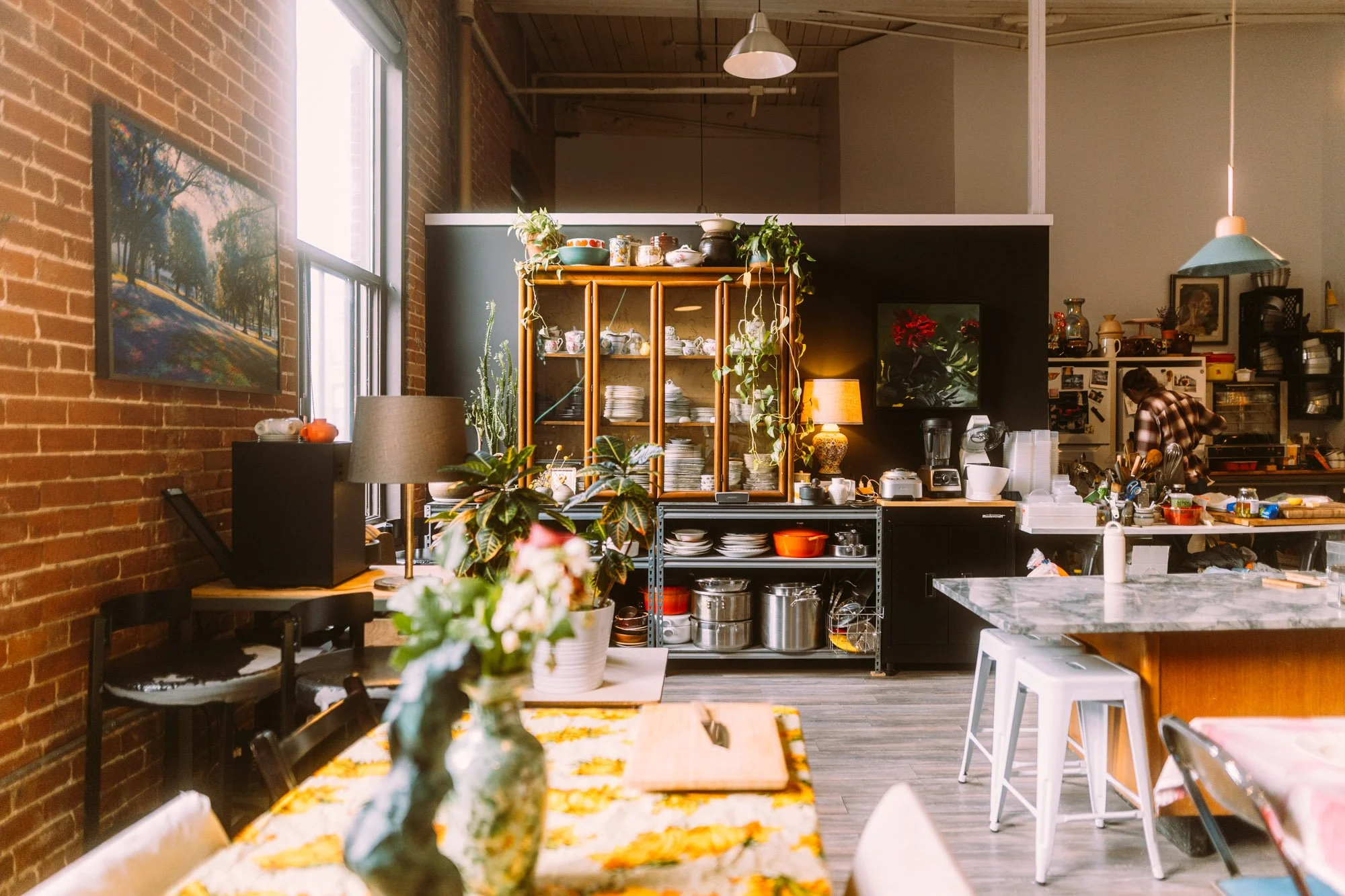 A cozy kitchen with exposed brick walls, large window, and a variety of plants. There is a wooden shelf with dishes, a small table with a floral tablecloth, and kitchen appliances like a blender and toaster. A person in a plaid shirt is working in the background.
