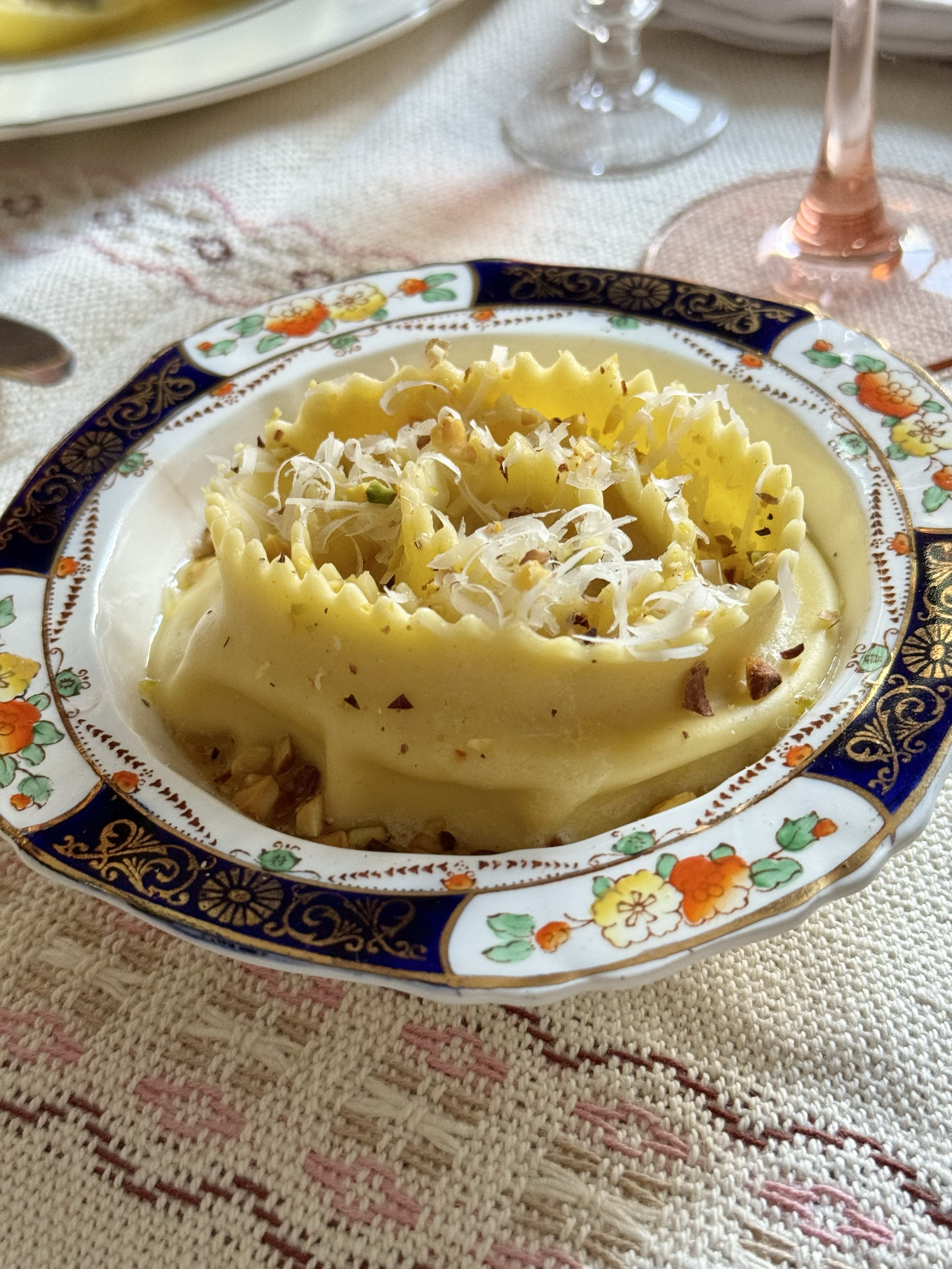 A plate of ravioli pasta topped with grated cheese and chopped nuts on an ornate floral-patterned plate, set on a tablecloth with glassware in the background.