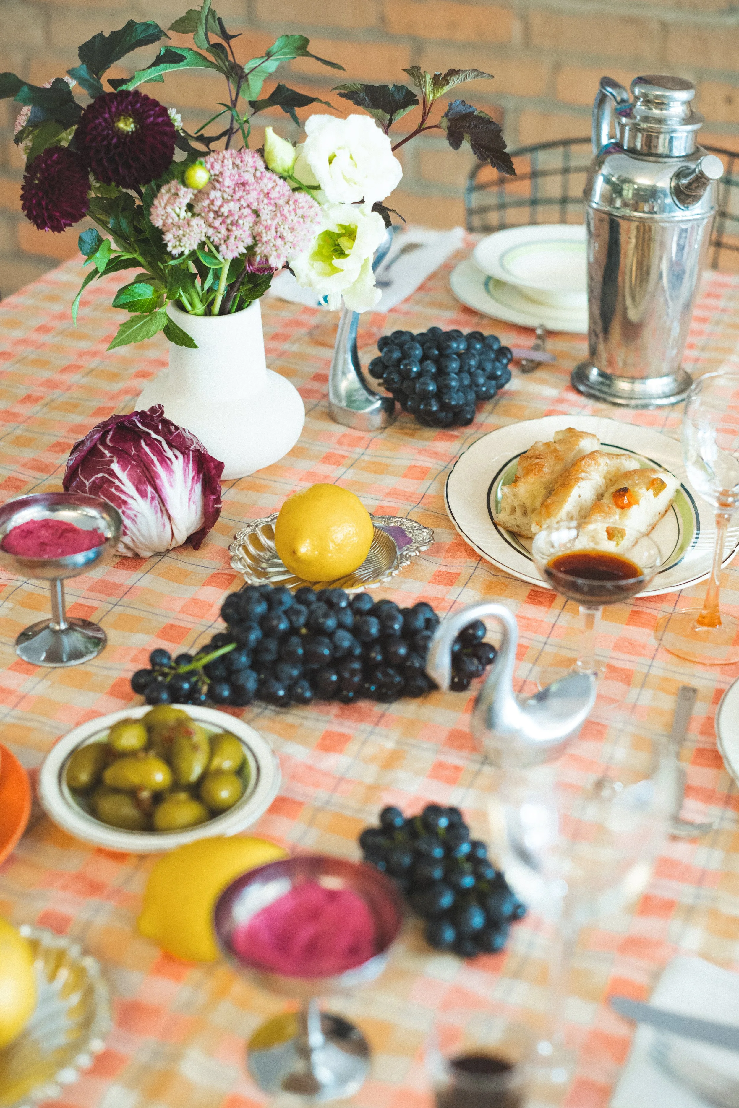 A table set for a meal with a checkered tablecloth, a vase of mixed flowers, plates, glasses, a pitcher, grapes, olives, a lemon, a radicchio, and bread, with decorative swan-shaped napkin holders.