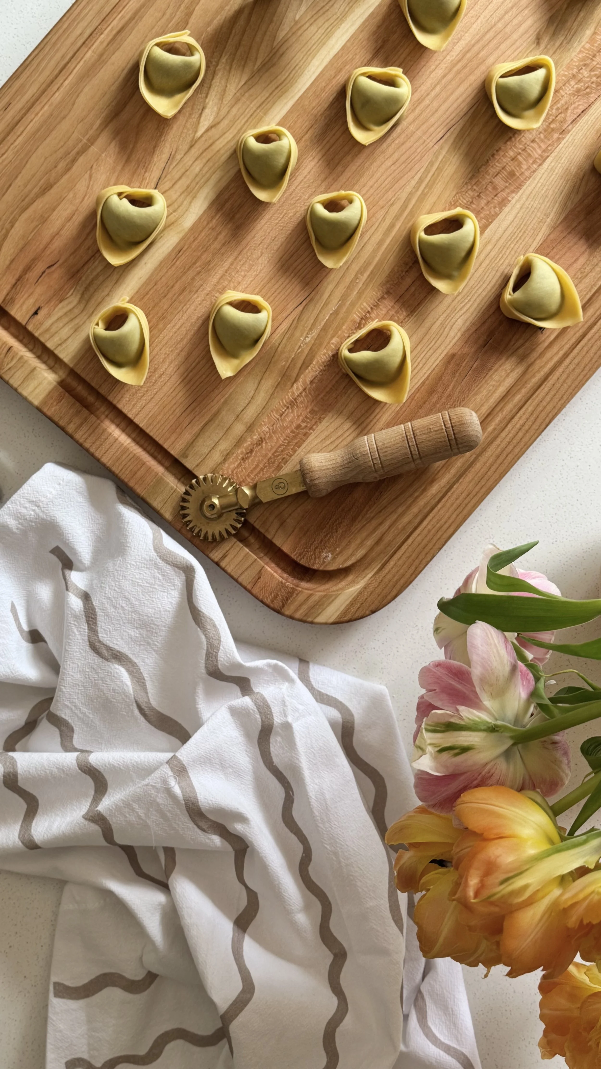 Tortellini vert sur une planche à découper en bois avec un coupe-pâte en bois, des fleurs roses et jaunes dans un vase et un torchon blanc avec des rayures beige sur une surface blanche.