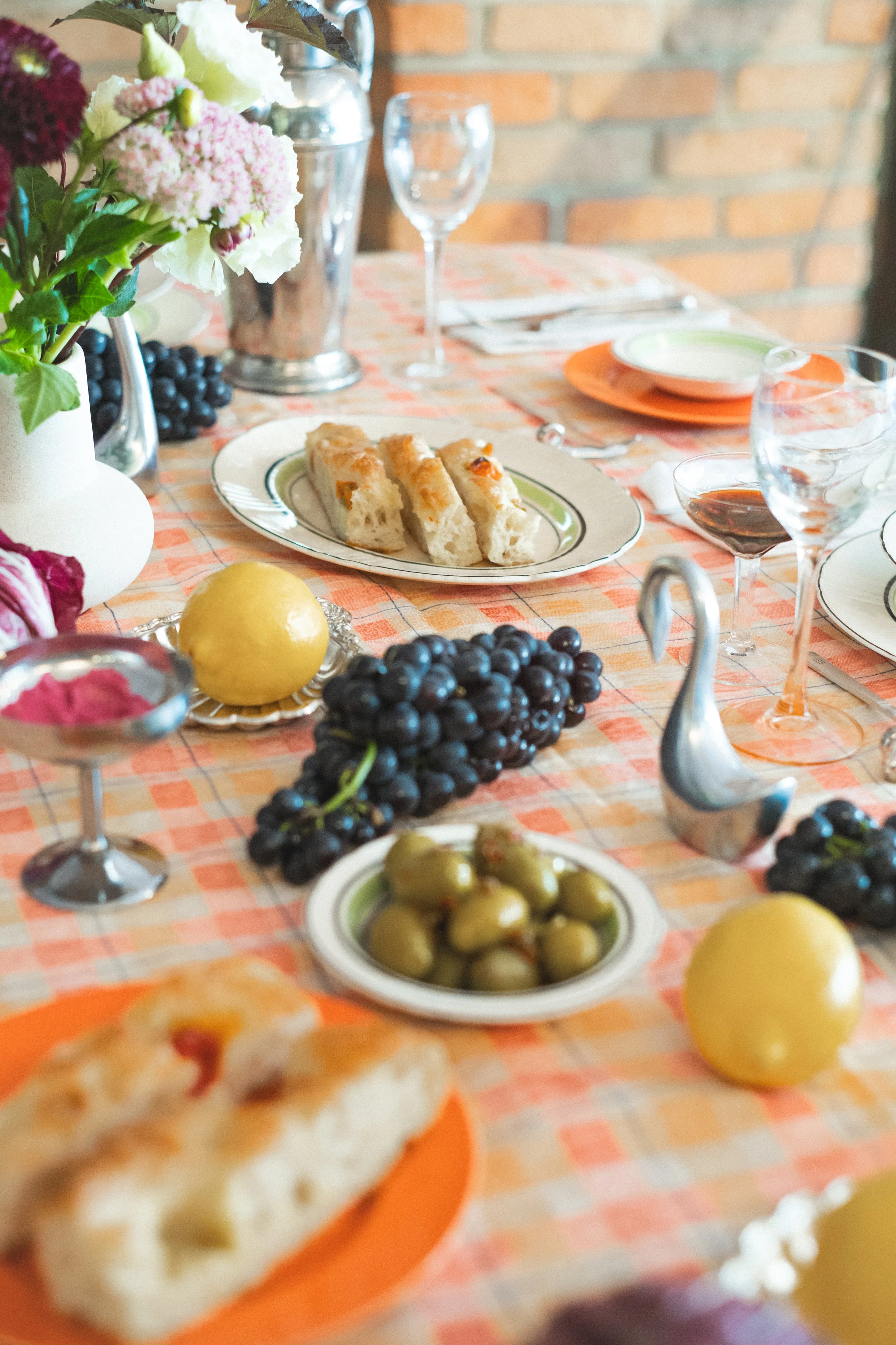 A table set for a meal with a checkered tablecloth, decorated with flowers, grapes, olives, lemon, bread, and glasses of wine.