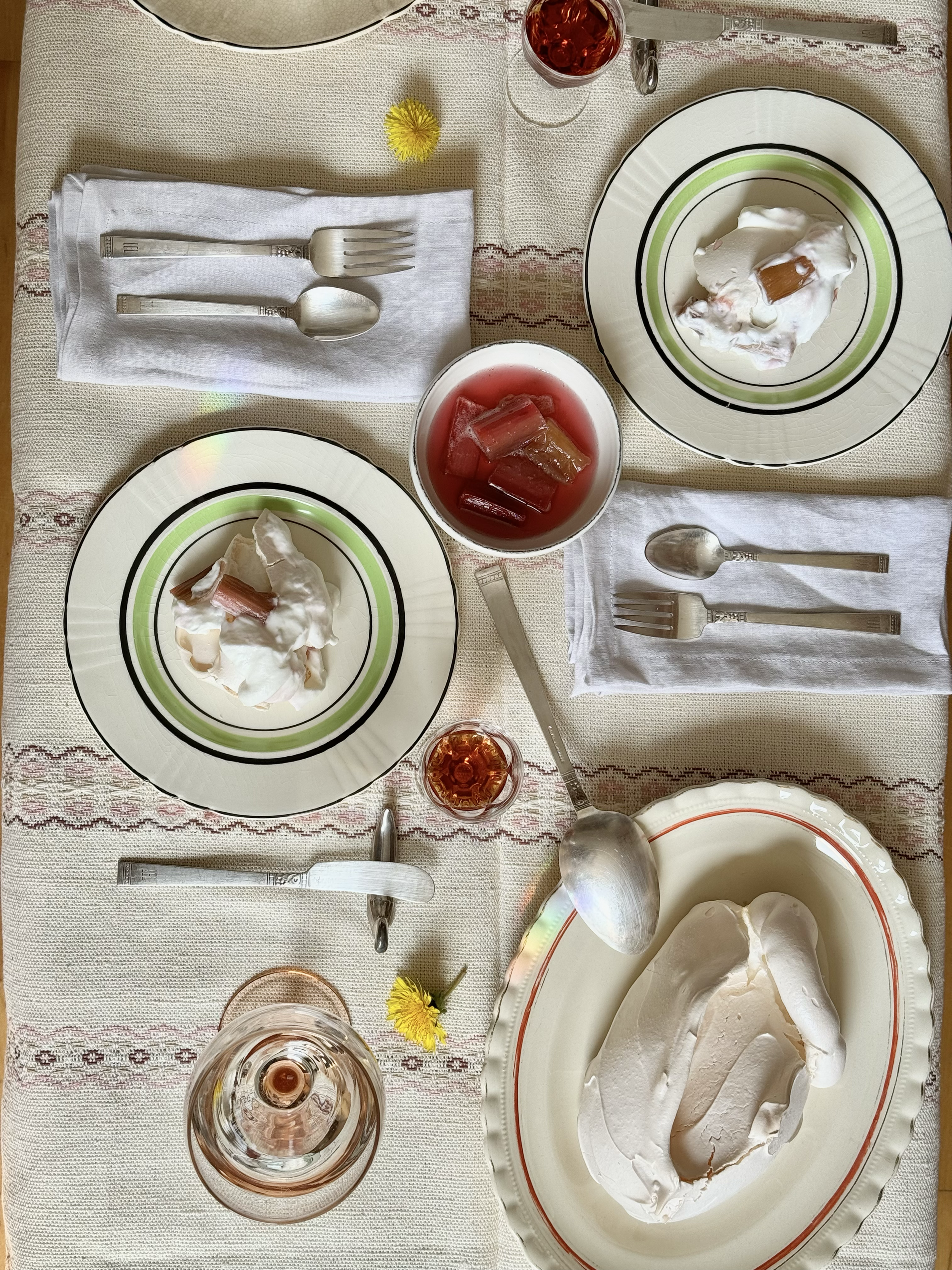 A family-style dessert table with plates of whipped cream topped with rhubarb, a bowl of pink-colored rhubarb compote, glasses of iced rhubarb juice, and silverware arranged on white cloth napkins on a cream-colored tablecloth decorated with embroide