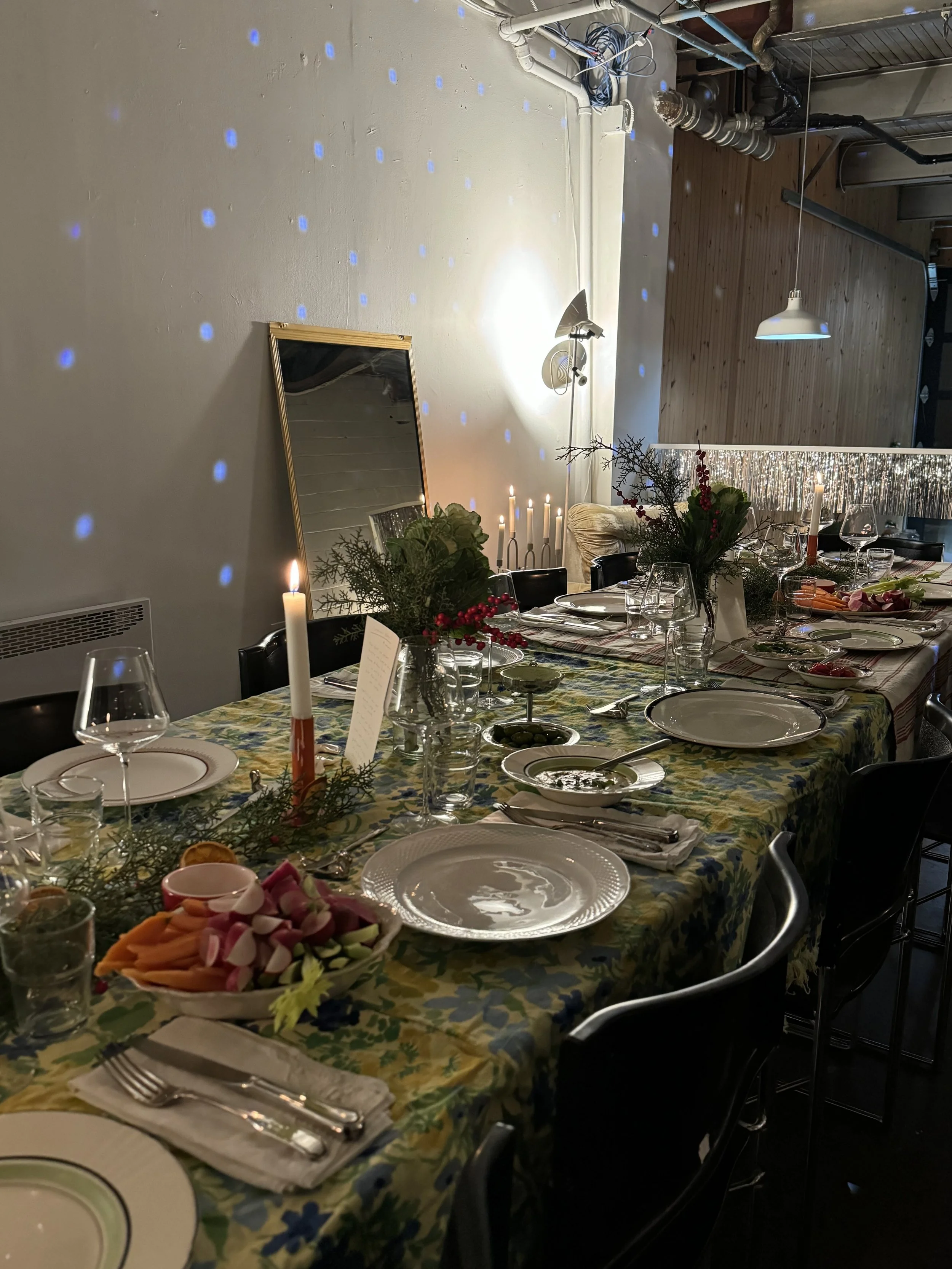 Indoor dining table decorated for a celebration with candles, floral arrangements, and plates, set with utensils and glasses, with a mirror and string lights on the wall.