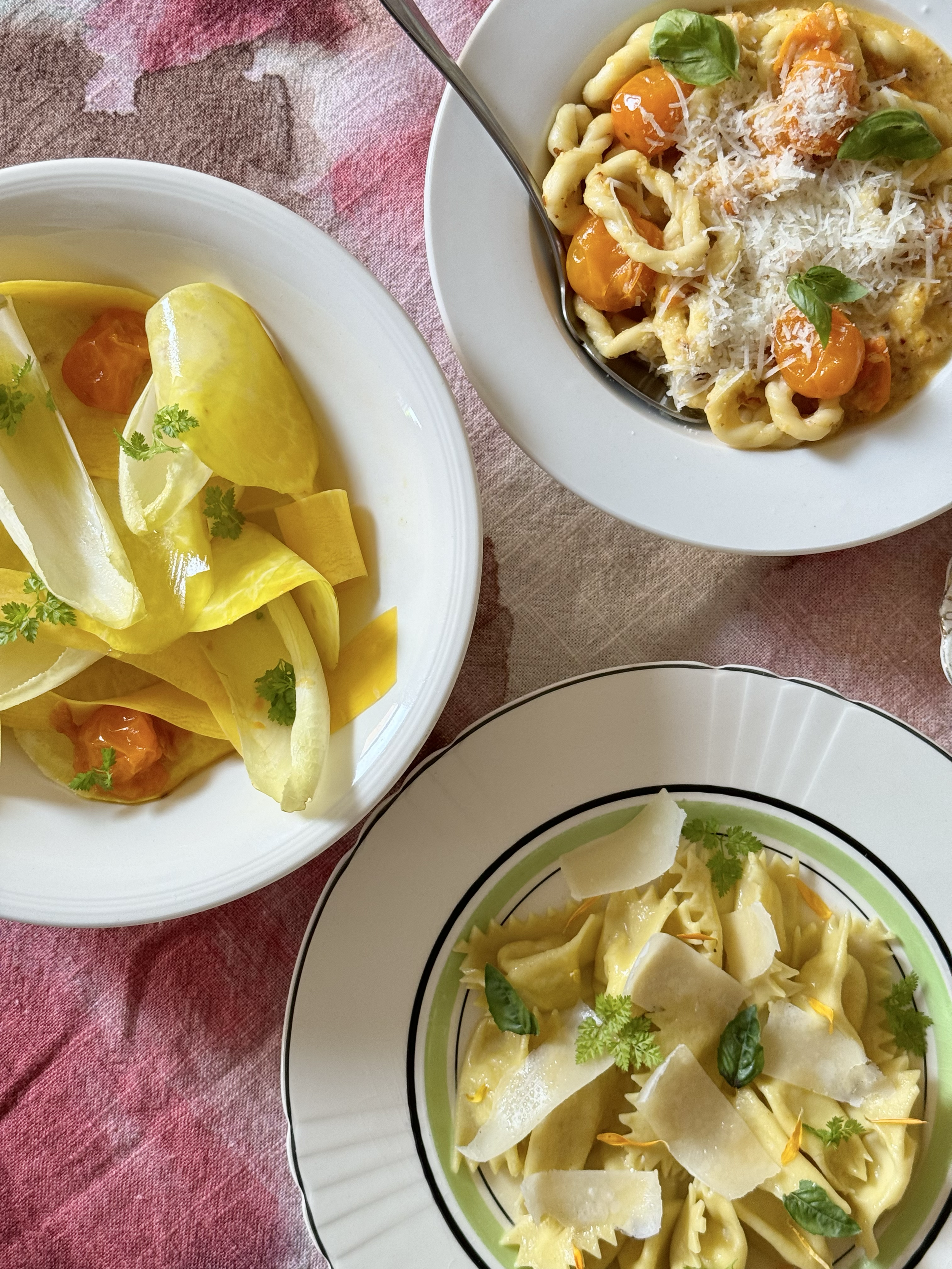 Three plates of pasta and a bowl of yellow lemon wedges with cherry tomatoes and fresh herbs on a pink tablecloth.