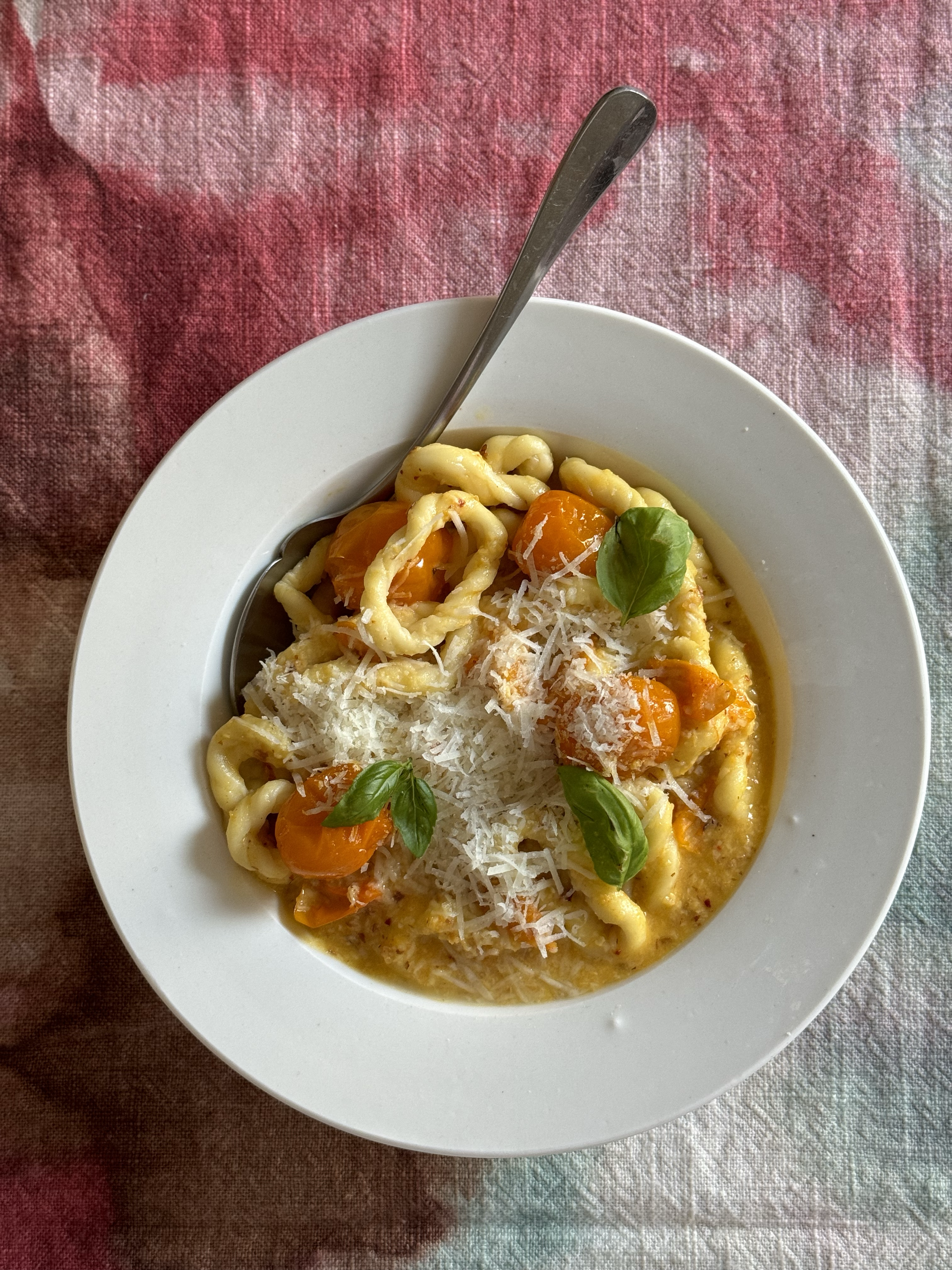 Plate of pasta with cherry tomatoes, grated cheese, and basil leaves on a multicolored fabric tablecloth.