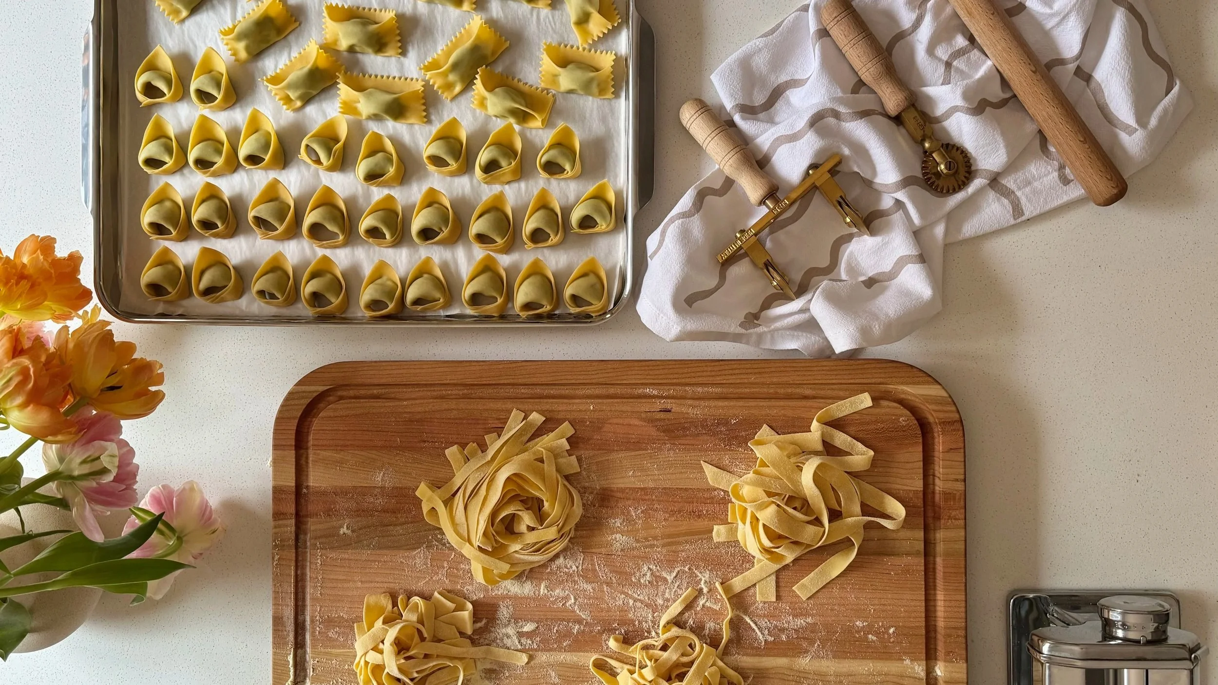 Pâtes fraîches en forme de tortellini prêtes à cuire, avec des pâtes coupées en rubans sur une planche en bois, une plaque de cuisson avec des tortellini non cuits, et divers outils de cuisine sur une surface blanche.