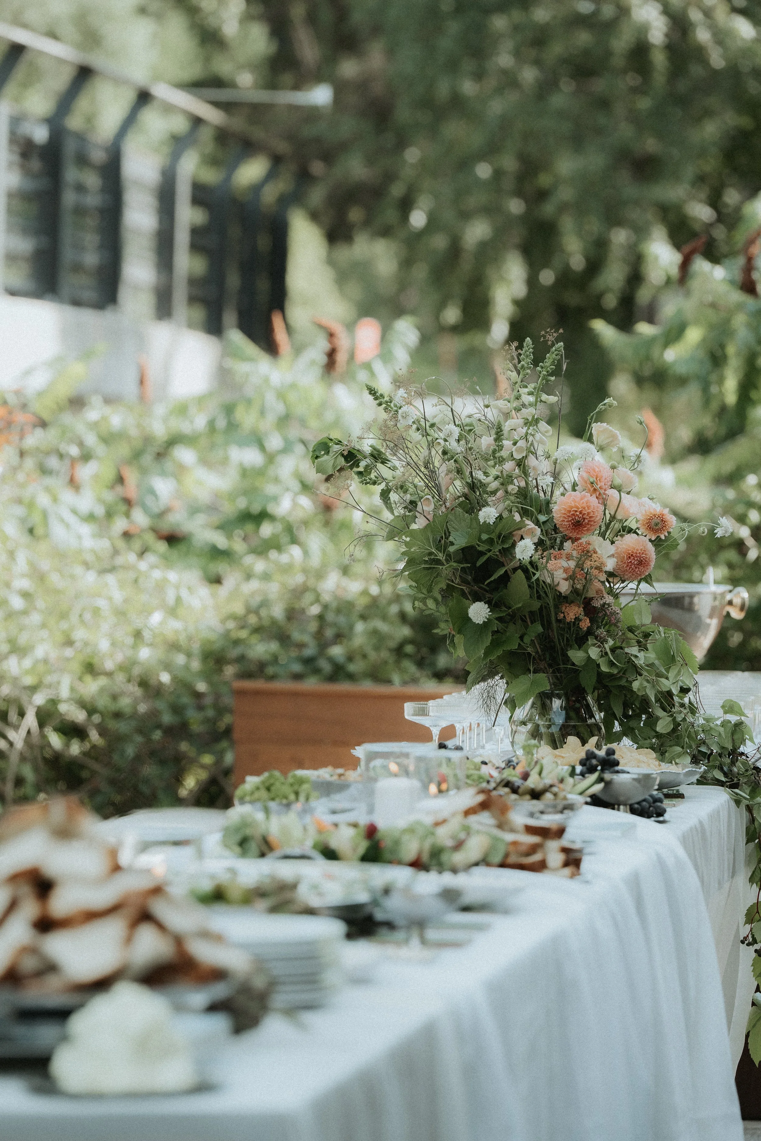 A long outdoor table decorated with a white tablecloth, topped with a large floral arrangement of pink and white flowers and greenery. The table is set with various appetizers, plates, and bowls, and is situated in a lush garden or backyard area.