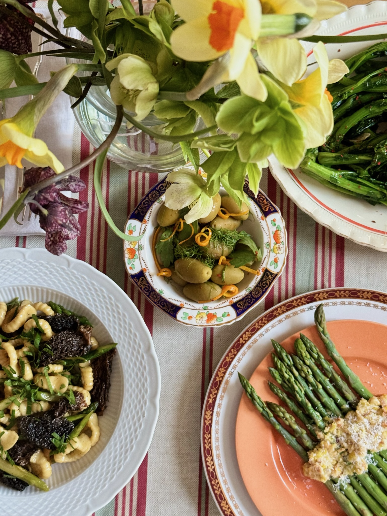 A table setting with a bouquet of yellow and purple daffodils and four plates of food: a bowl of marinated green beans, a plate of cooked asparagus topped with grated cheese, a bowl of marinated small pickles with herbs and citrus peel, and a plate o