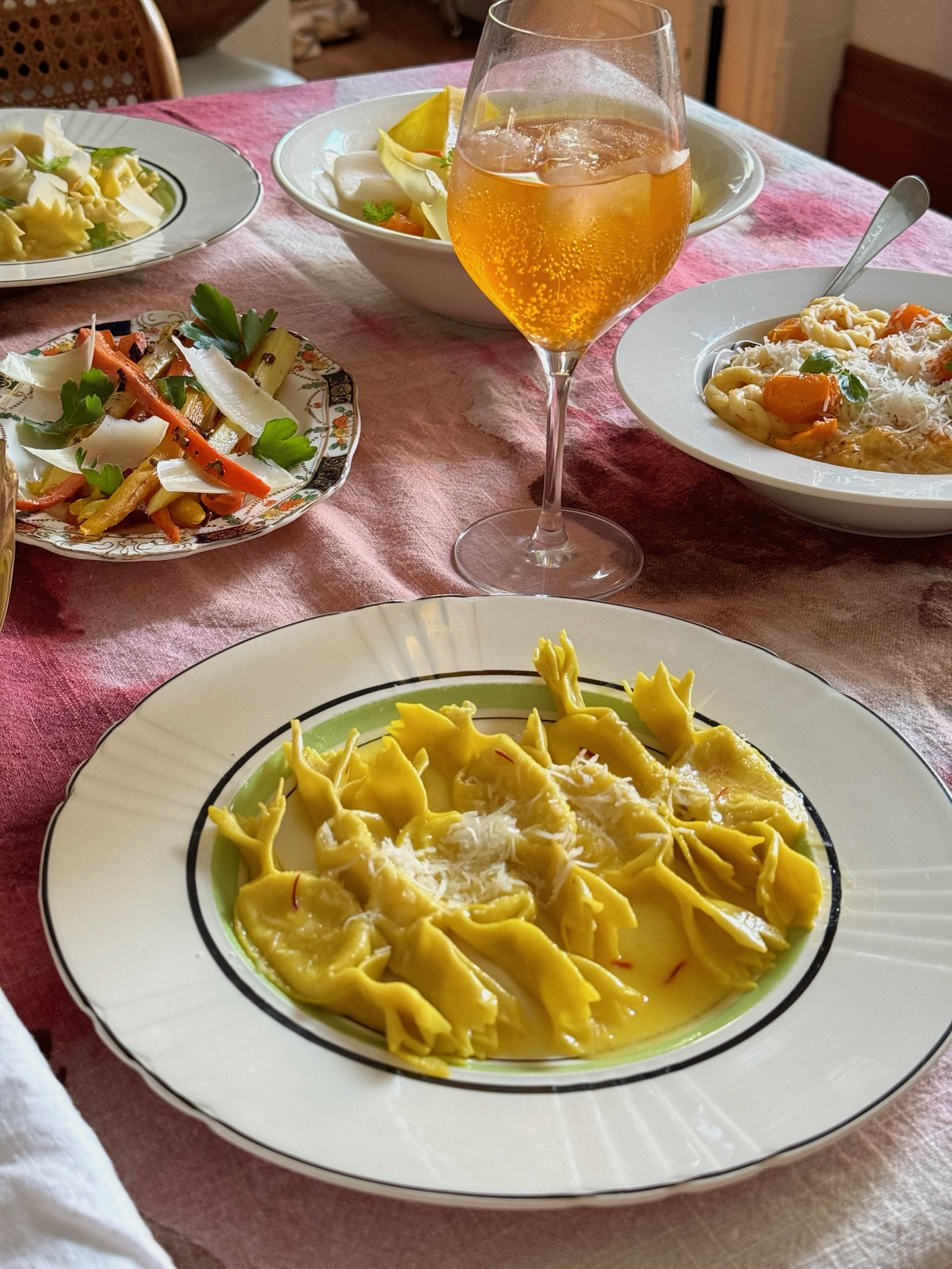 A dinner table with various dishes including tortellini pasta with grated cheese, a bowl of salad, another bowl of pasta with cherry tomatoes, a plate with vegetable stir-fry, and a glass of sparkling golden beverage on a pink tablecloth.