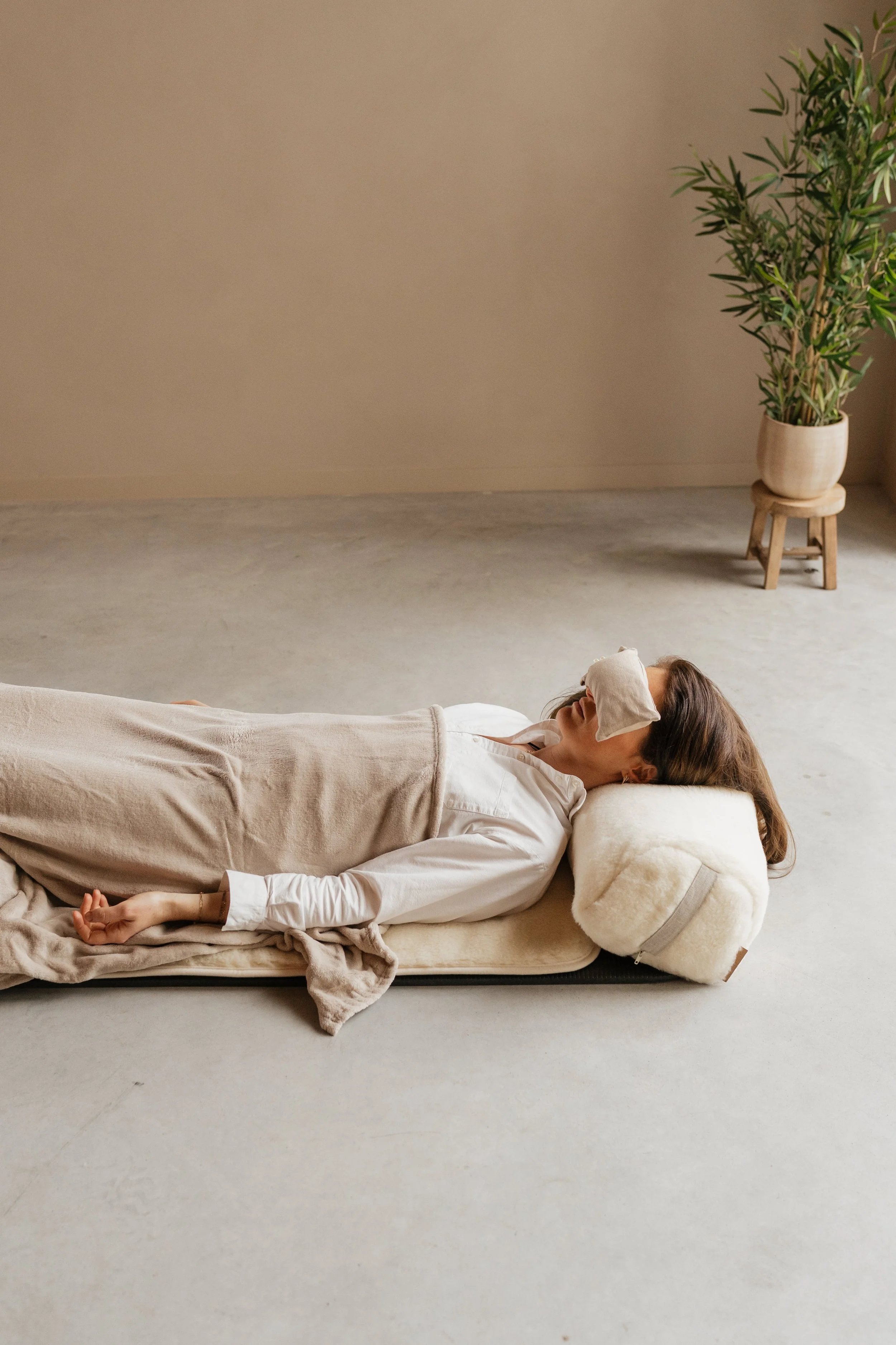 A woman lying on her side on a bed, with an eye mask over her eyes, resting on a comfortable pillow, in a minimalistic room with neutral colors and a potted plant in the background.
