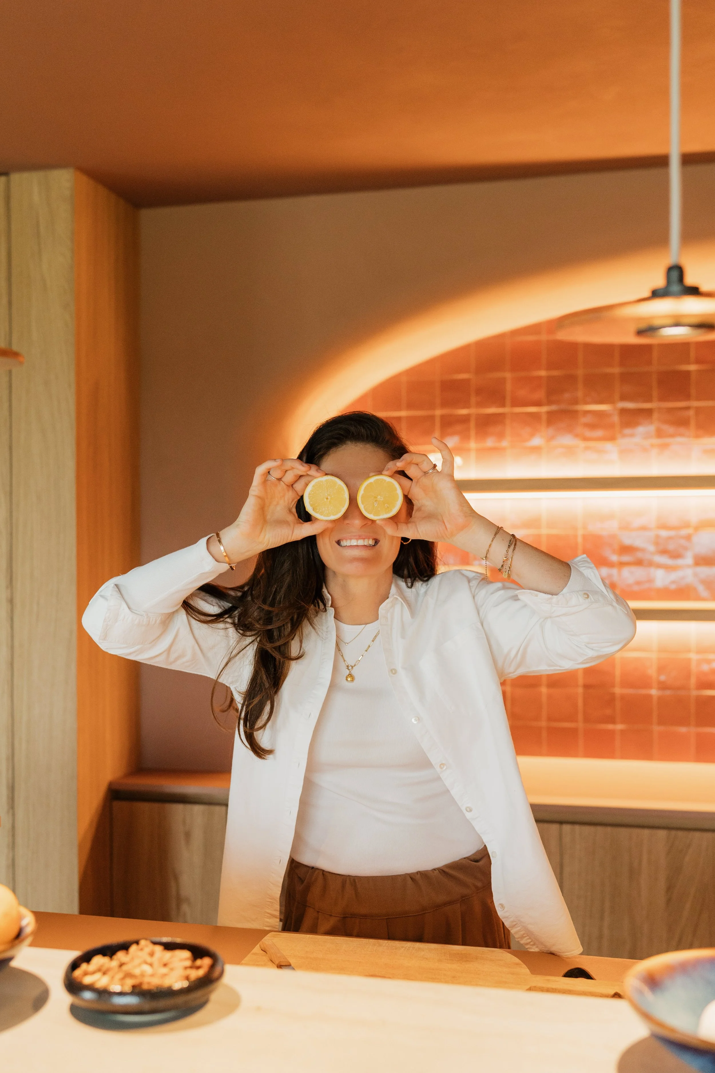 A woman smiling and holding two lemon halves in front of her eyes inside a kitchen with warm lighting and wooden accents.
