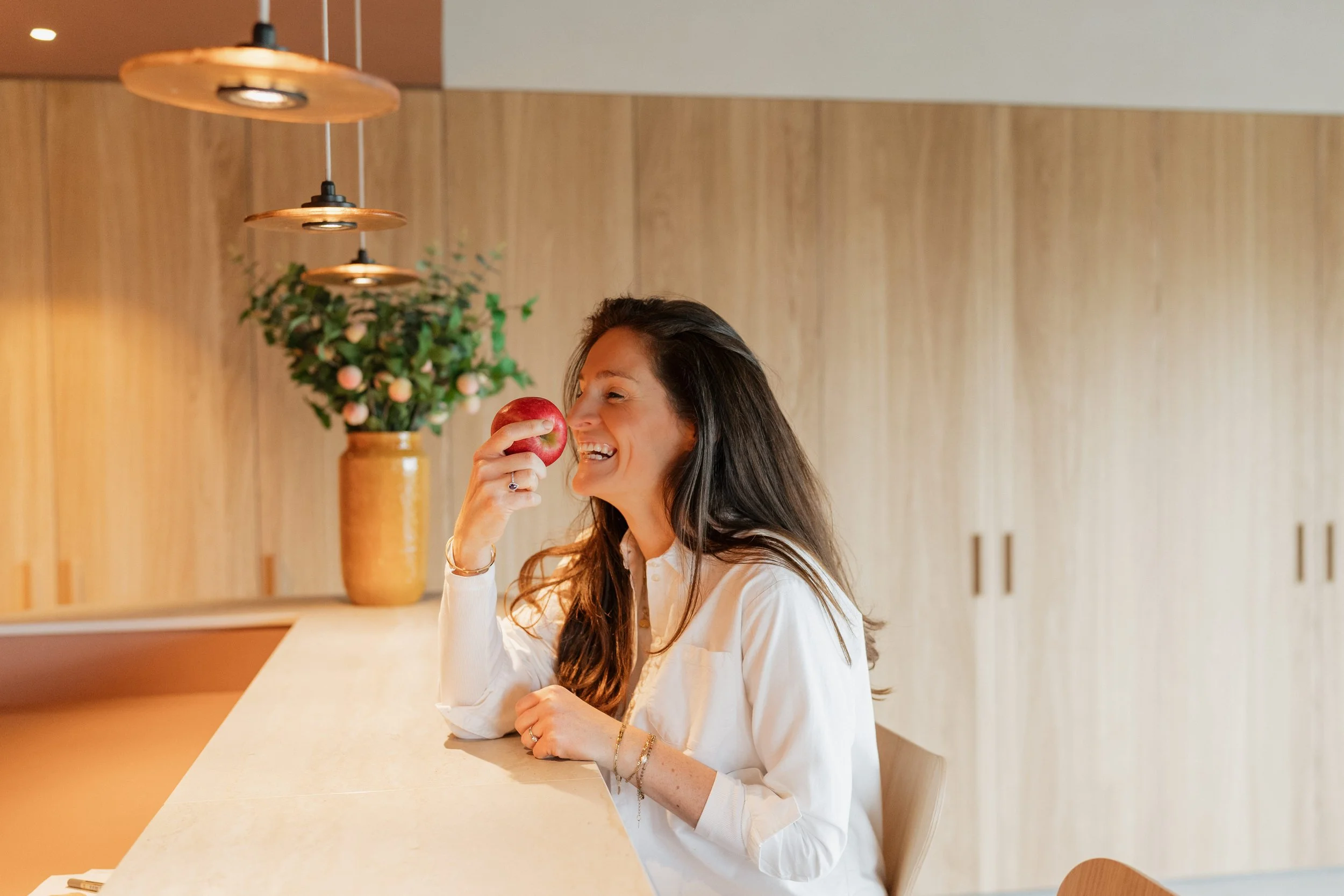 A woman with long dark hair smiling while holding an apple near her nose while sitting at a kitchen counter with wooden cabinets and a vase with plants in the background.