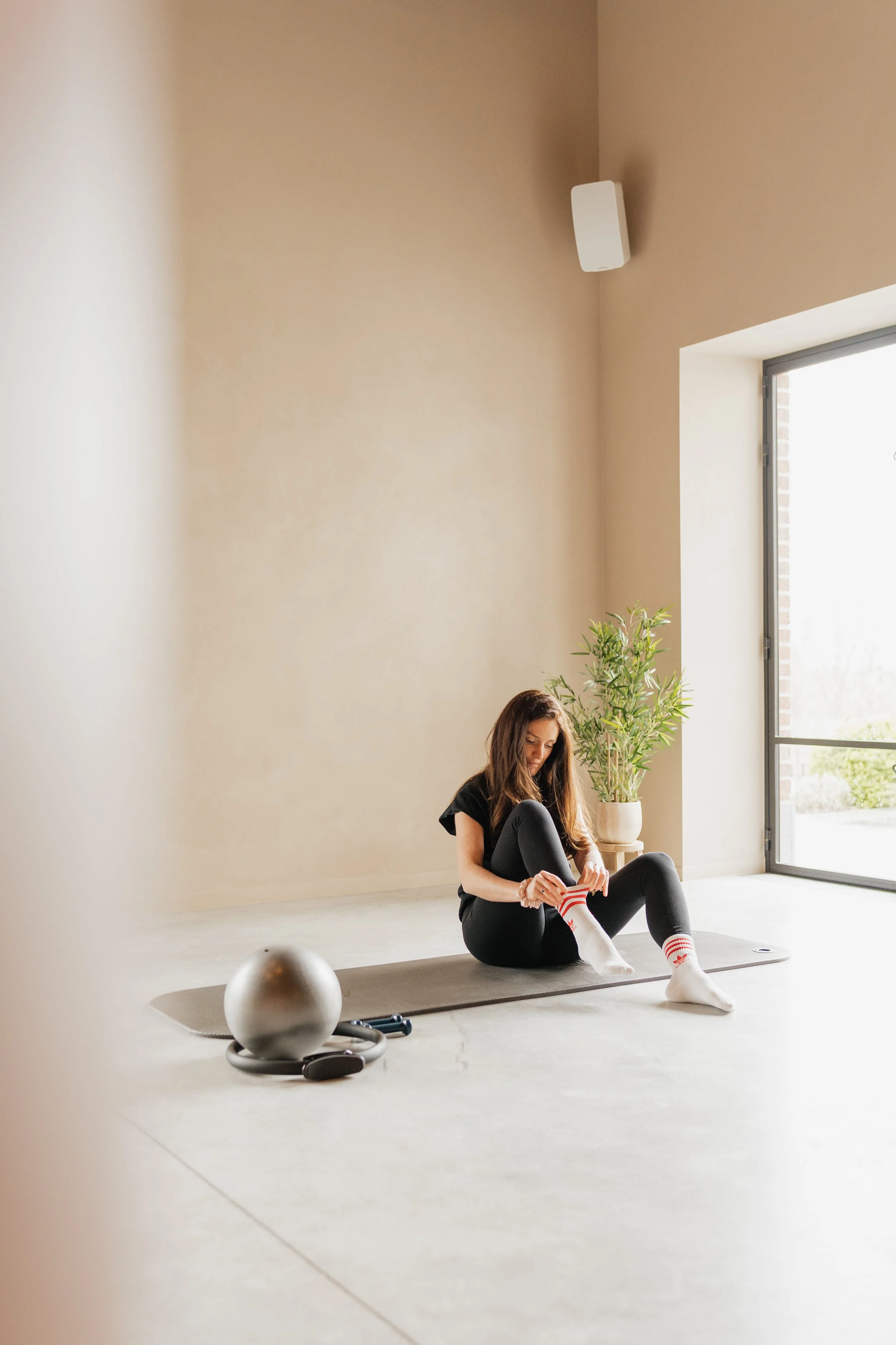 A woman sitting on a yoga mat in a brightly lit room, putting on striped socks. There is a large potted plant and a window in the background, with exercise equipment including a silver ball, jump rope, and dumbbell placed nearby.