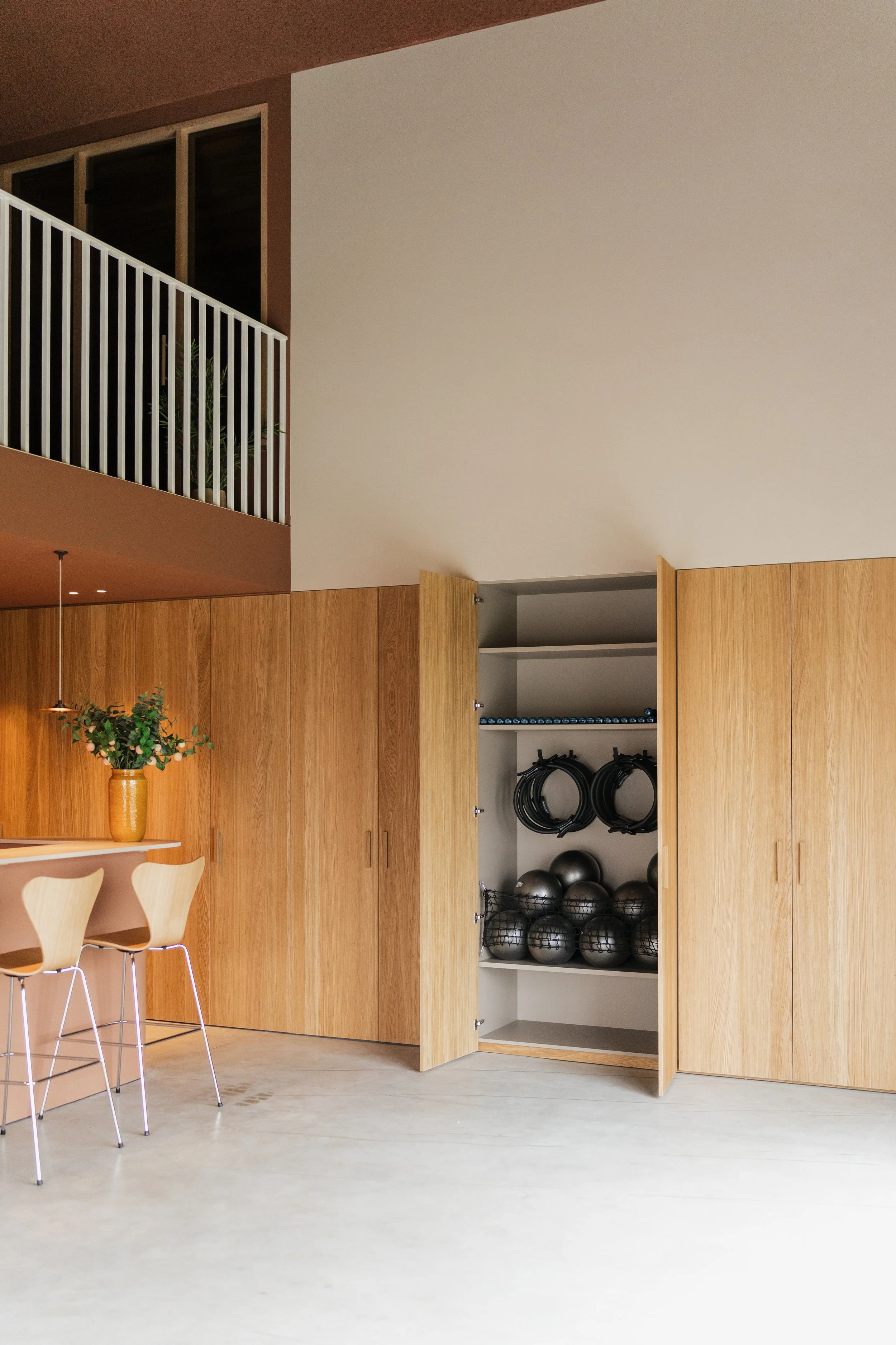 An indoor space with wooden cabinets, a white shelf with black and silver exercise balls, resistance bands, and medicine balls, and a partial view of a dining area with light-colored Fritz Hansen chairs and a vase with green foliage
