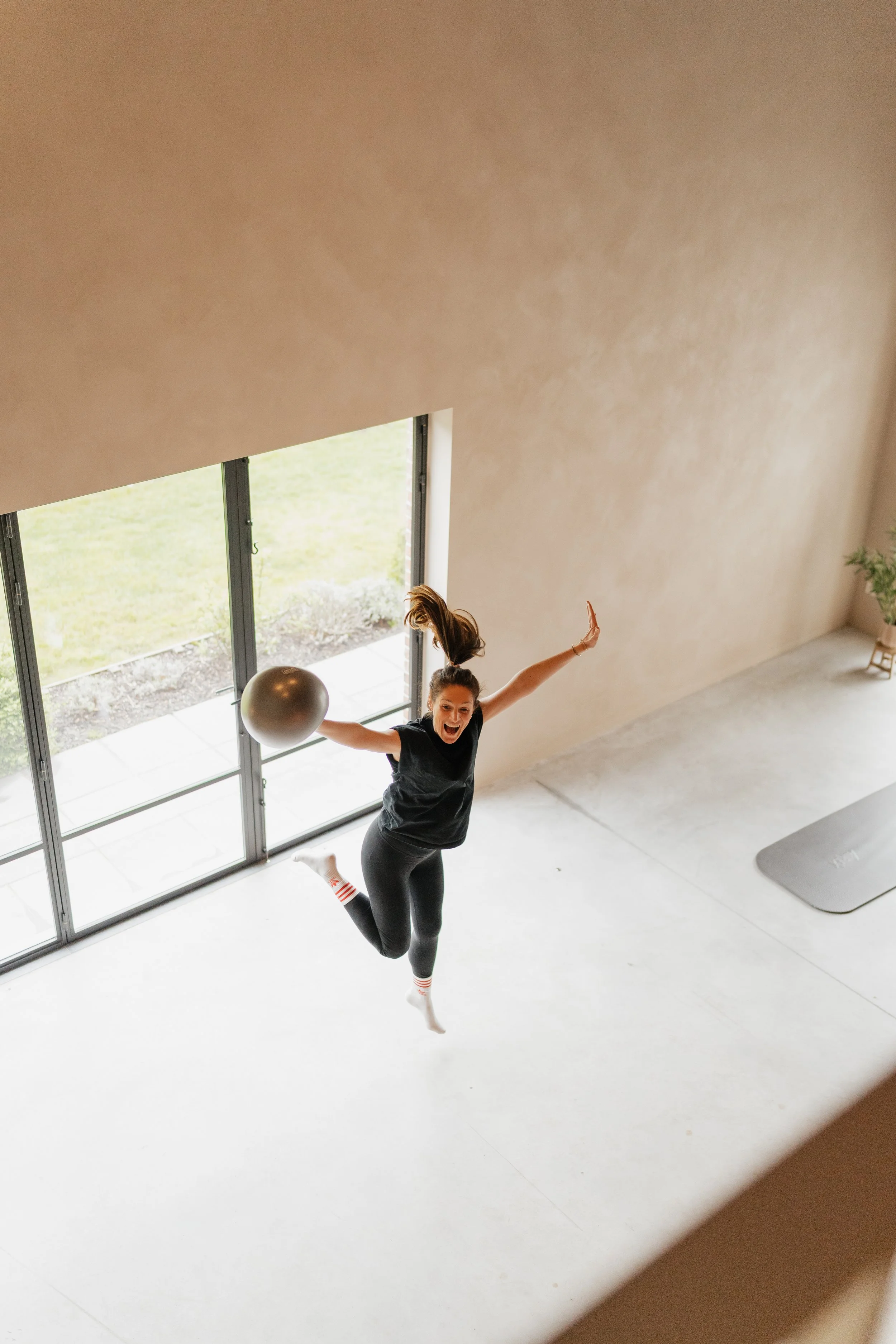 A woman in workout clothes jumping joyfully in a sunlit room, holding a large exercise ball.