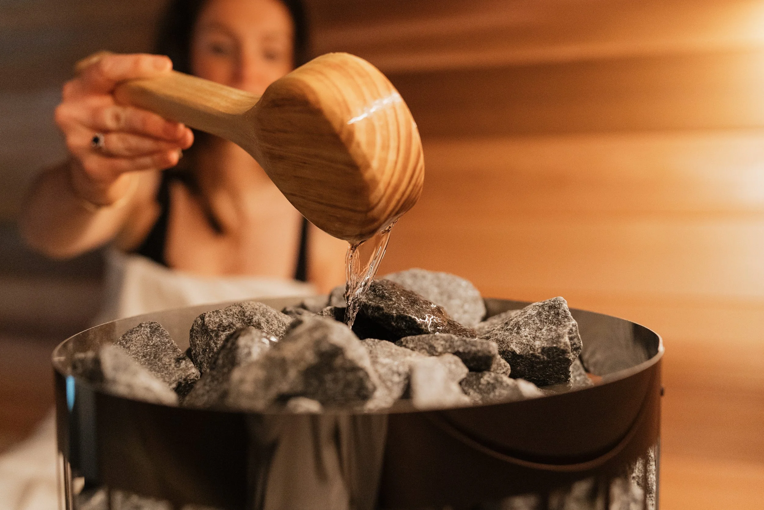 A woman is pouring water from a wooden ladle onto hot stones in a sauna.