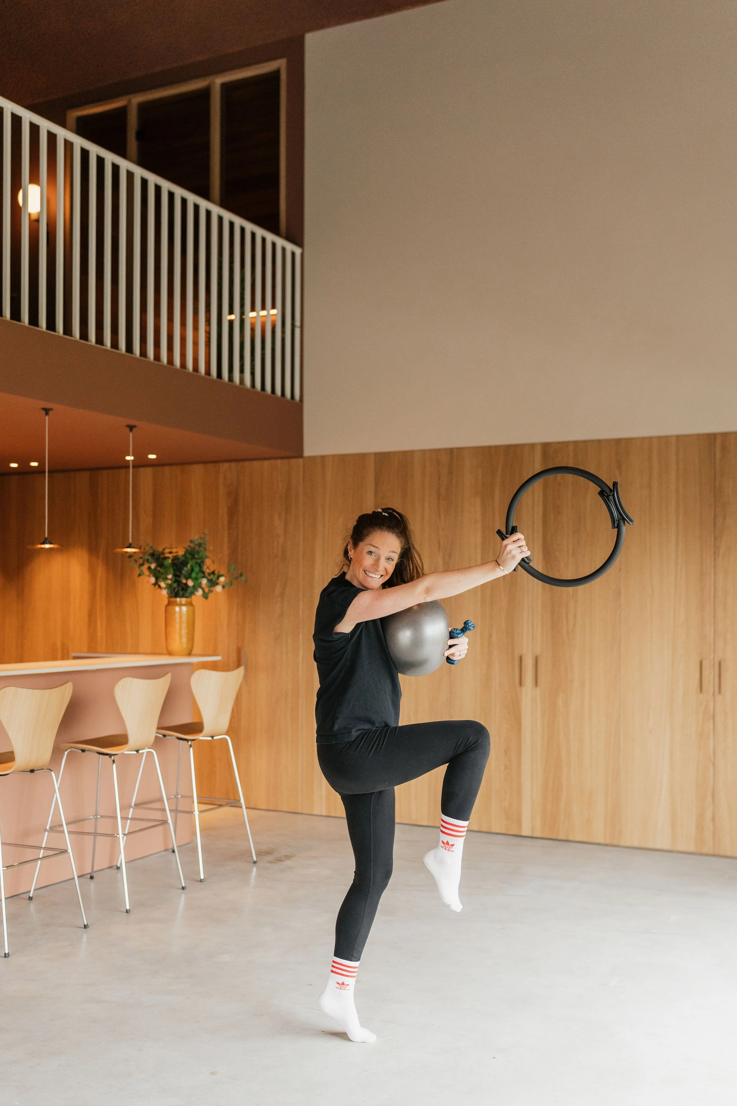 Woman in black workout clothes lifting a kettlebell and a suspension trainer in a modern, minimalistic room with wooden paneling and bar stools.
