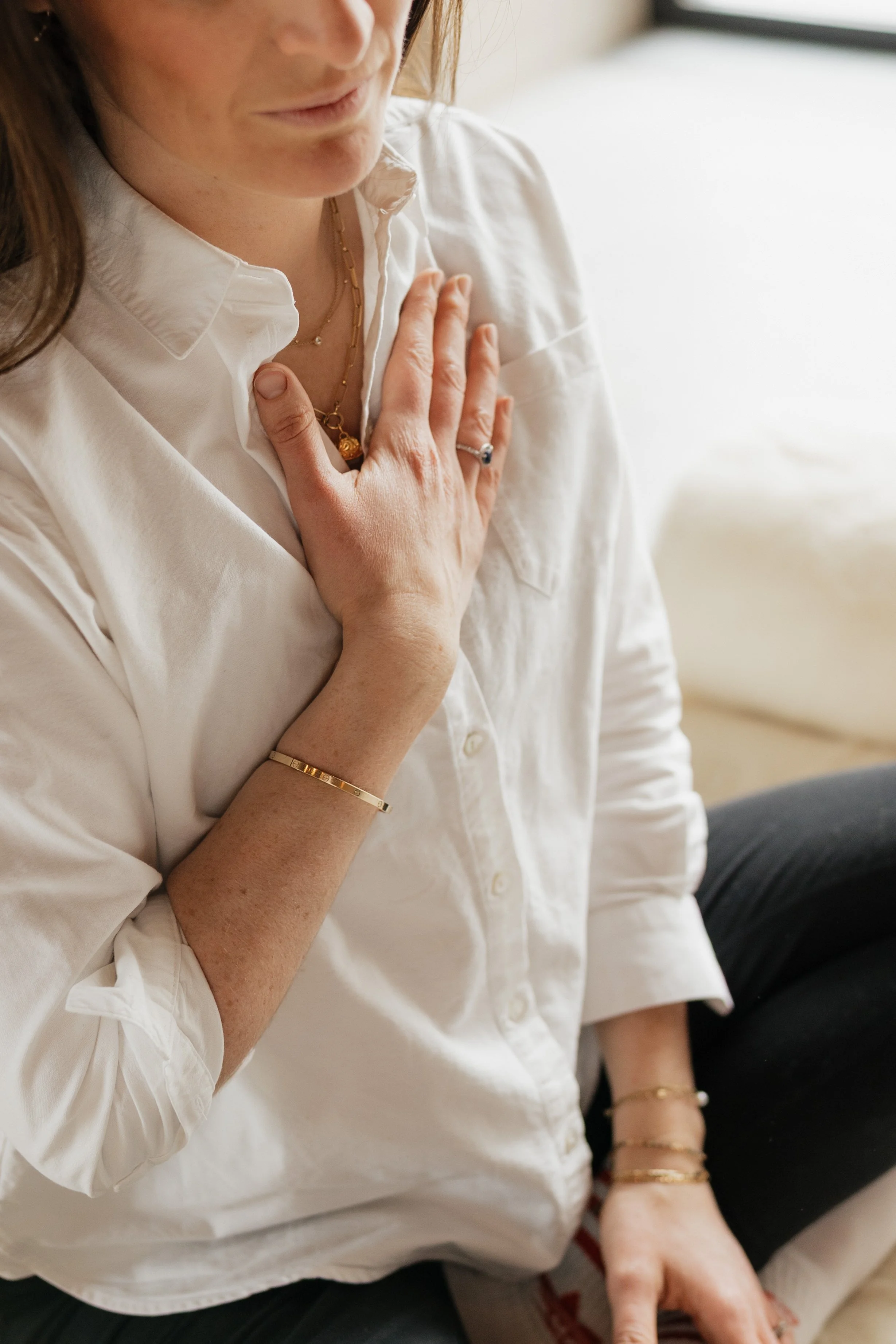 A woman wearing a white shirt, sitting on a couch, with her hand on her chest, showing gold jewelry including a bracelet, rings, and necklaces.